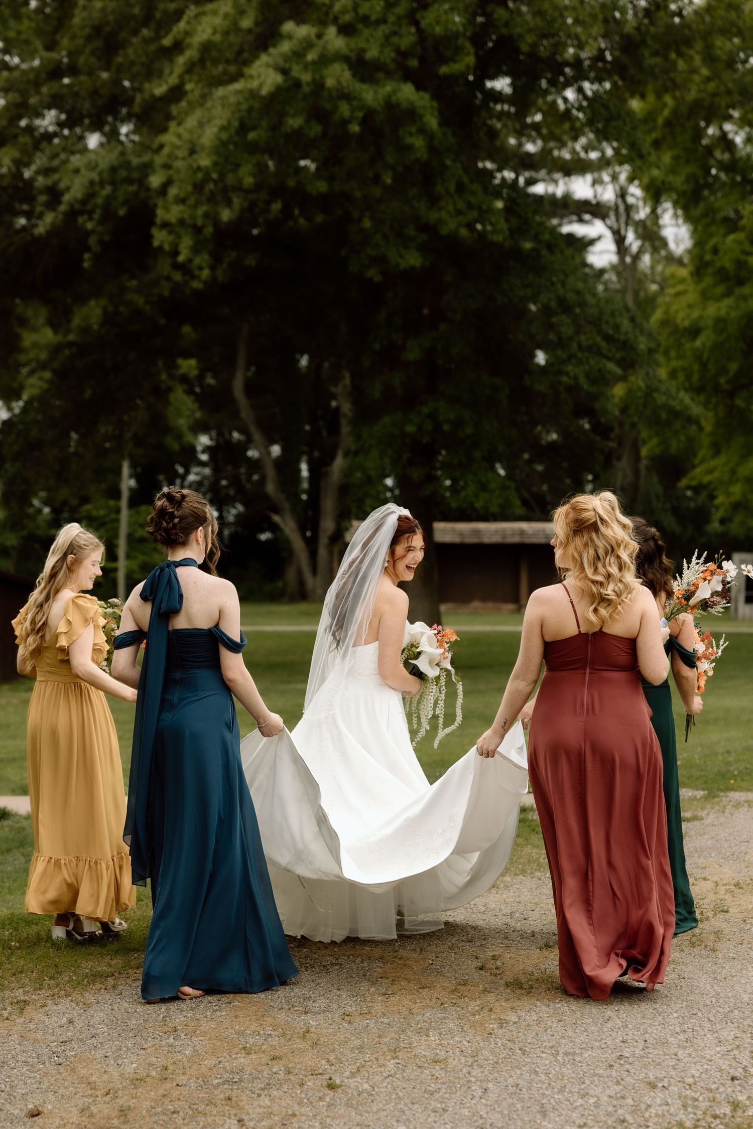 Back view of bride in white gown with veil surrounded by bridesmaids in colorful mismatched dresses.
