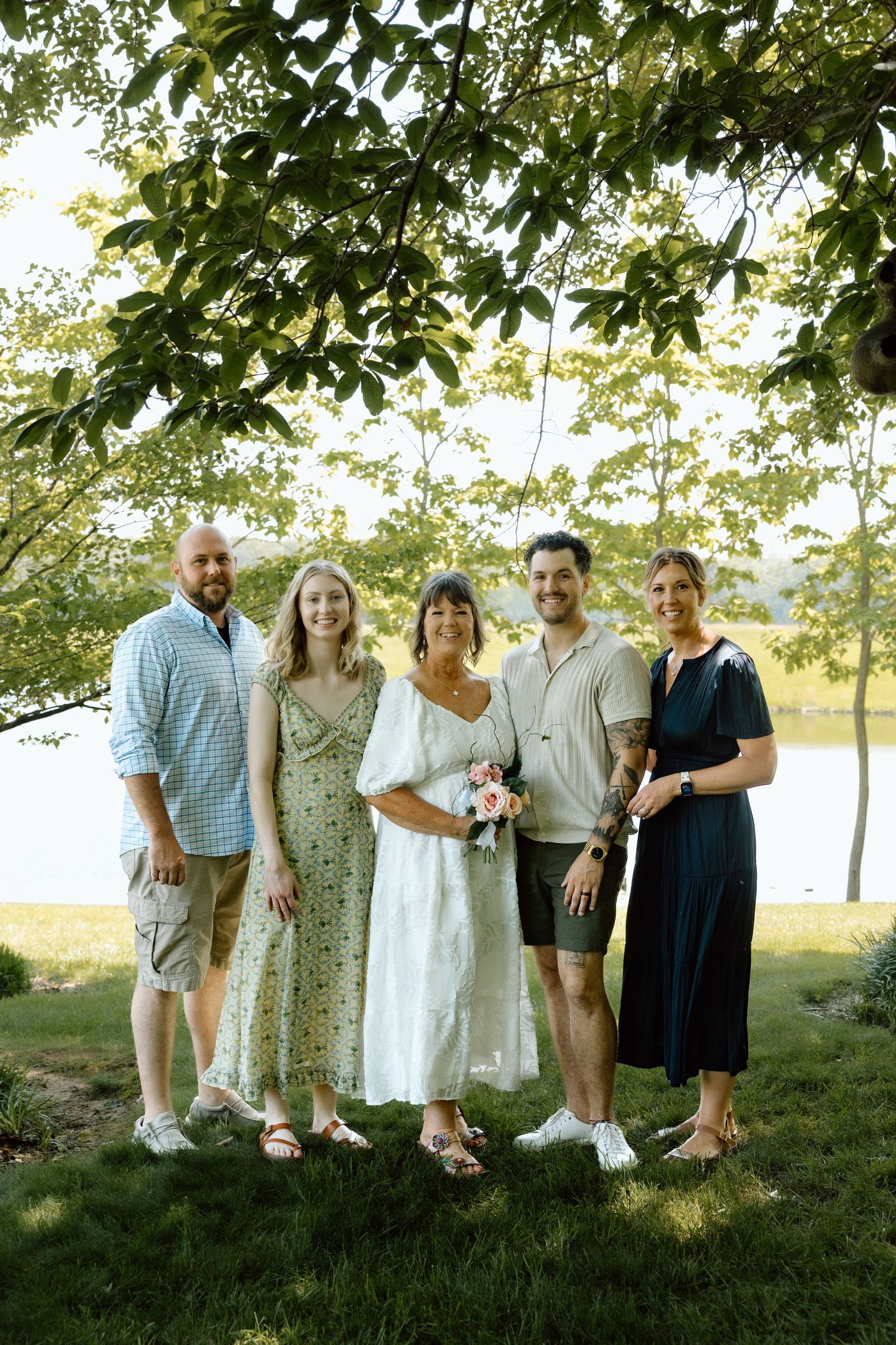 Bride with bouquet poses with four family members under leafy tree near lakeside after wedding.