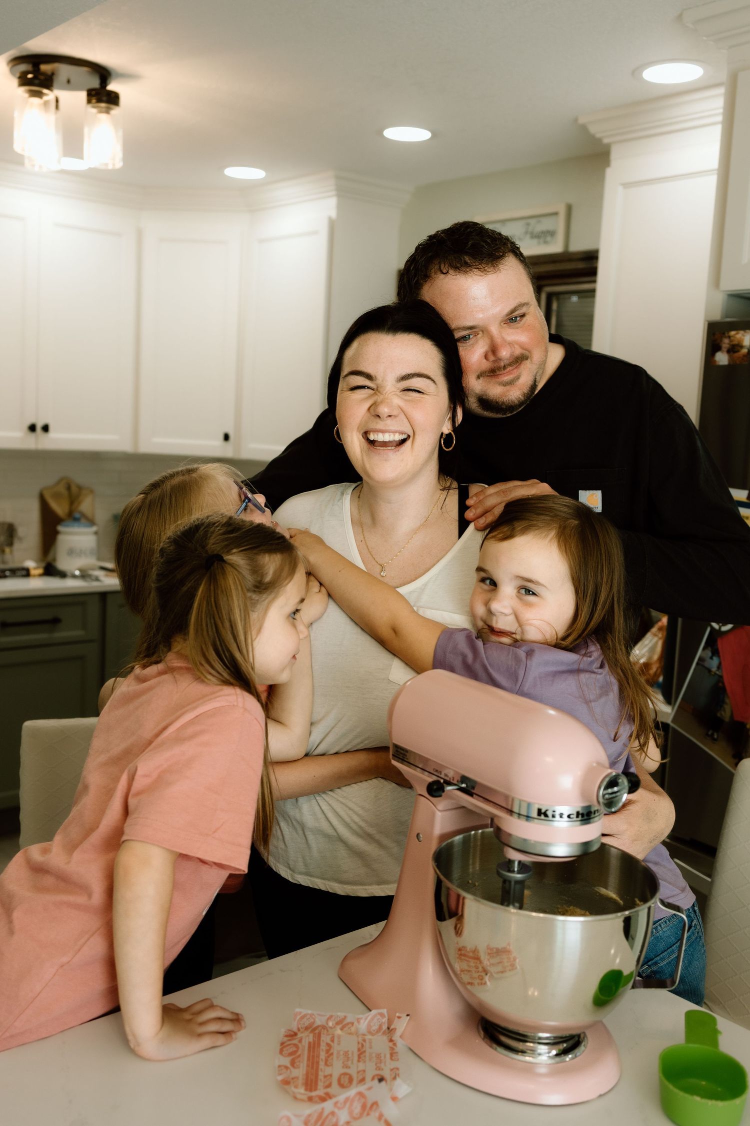Happy family laughing together around a kitchen island with a pink stand mixer during a baking session.