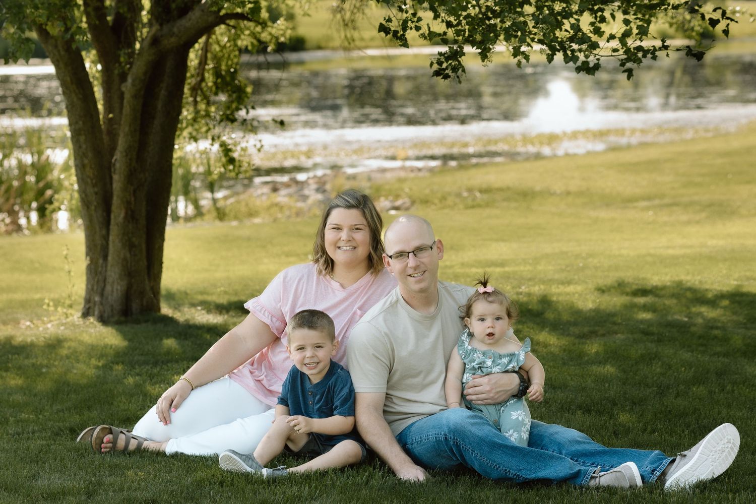 Happy family of four poses together on green grass under a tree with a peaceful river in the background.