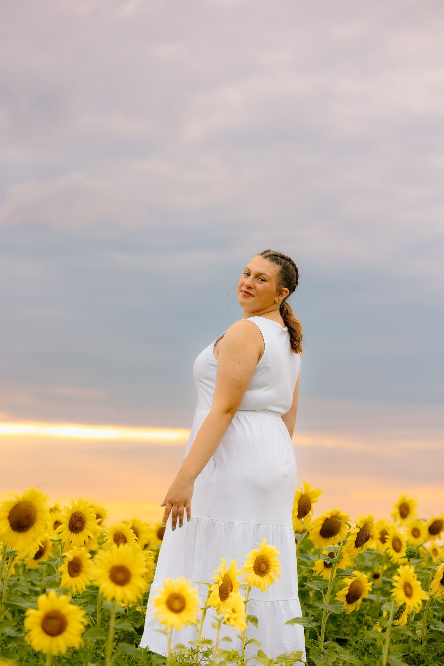 Woman in white dress posing gracefully in sunflower field beneath a dramatic pastel sunset sky.