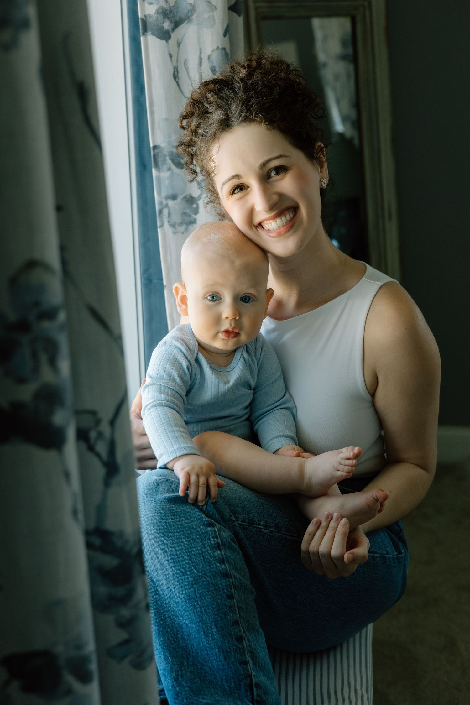Smiling young mother holding her baby in a white sleeveless top and jeans near a window with floral curtains.