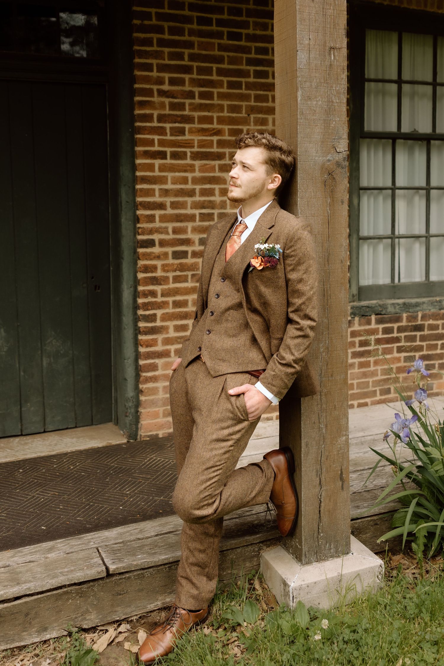 Dapper groom in brown tweed suit with boutonniere gazing upward while leaning against rustic wooden post.