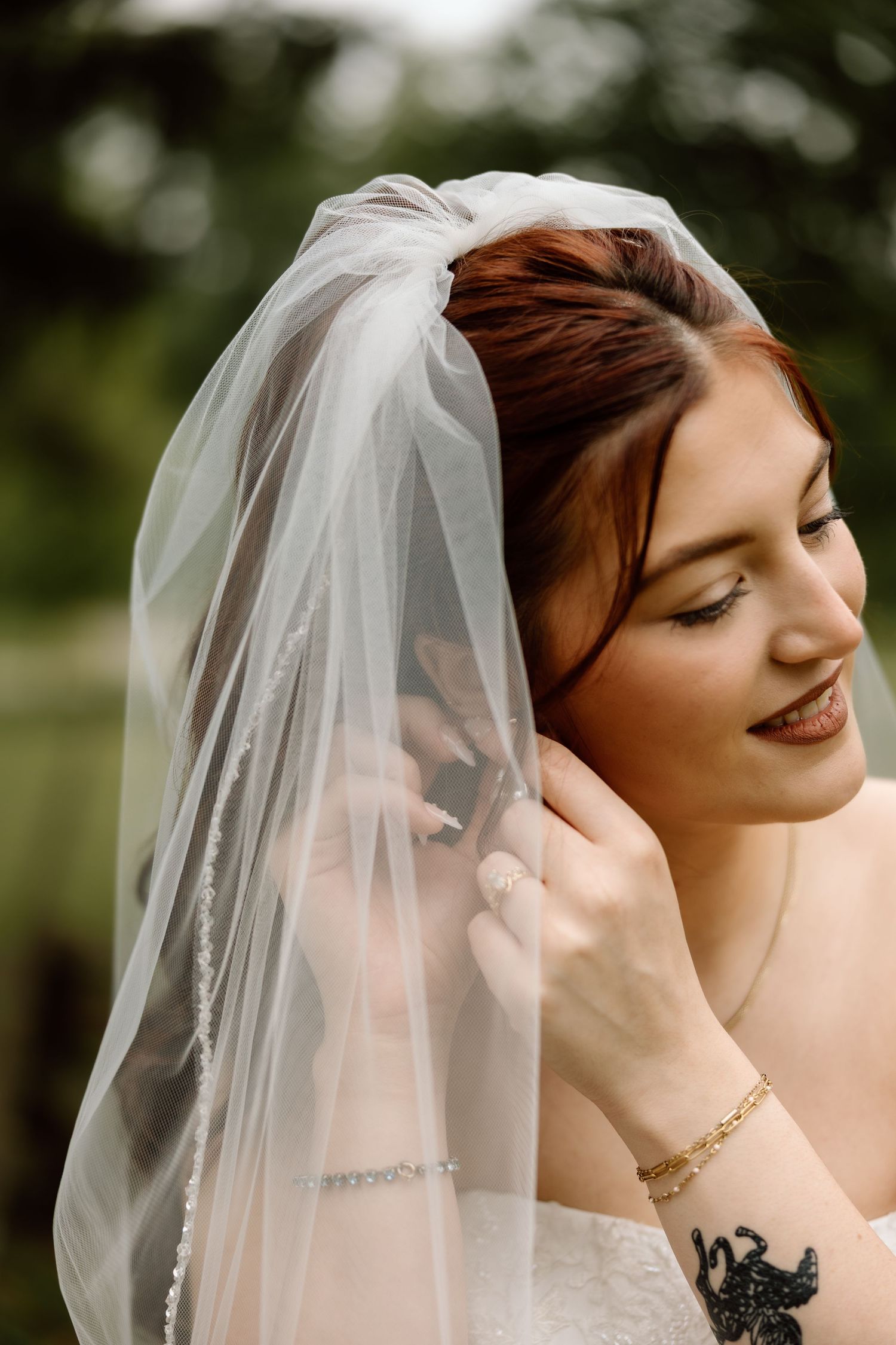 Radiant bride with auburn hair smiling softly while adjusting her delicate white veil outdoors.