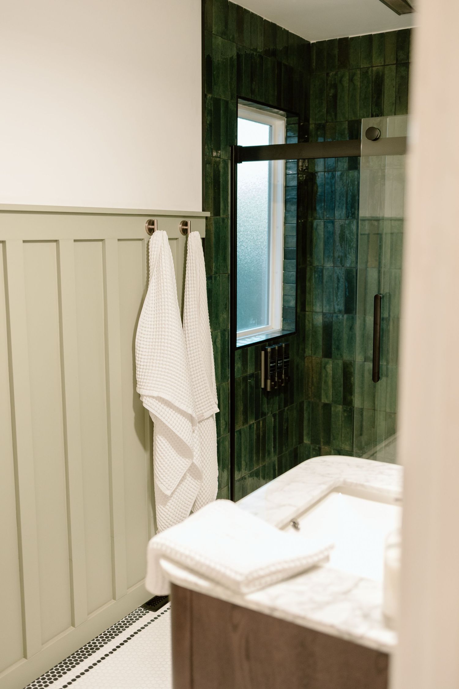Green zellige tile shower with glass door, wainscoting, and white towels on wooden vanity.