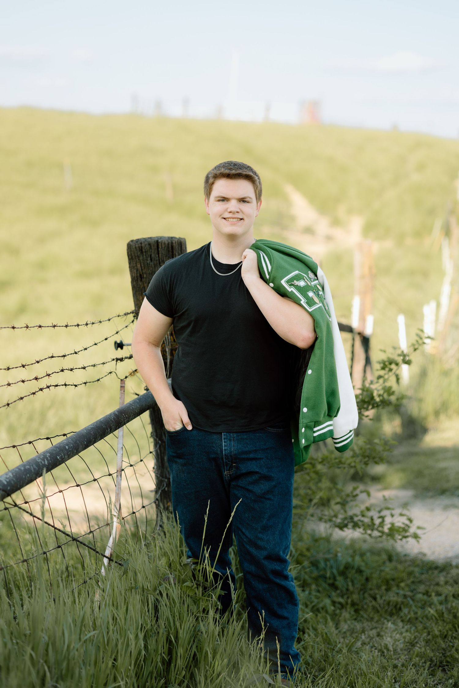 Young man in black tee and jeans holds green letterman jacket over shoulder by rural barbed wire fence.