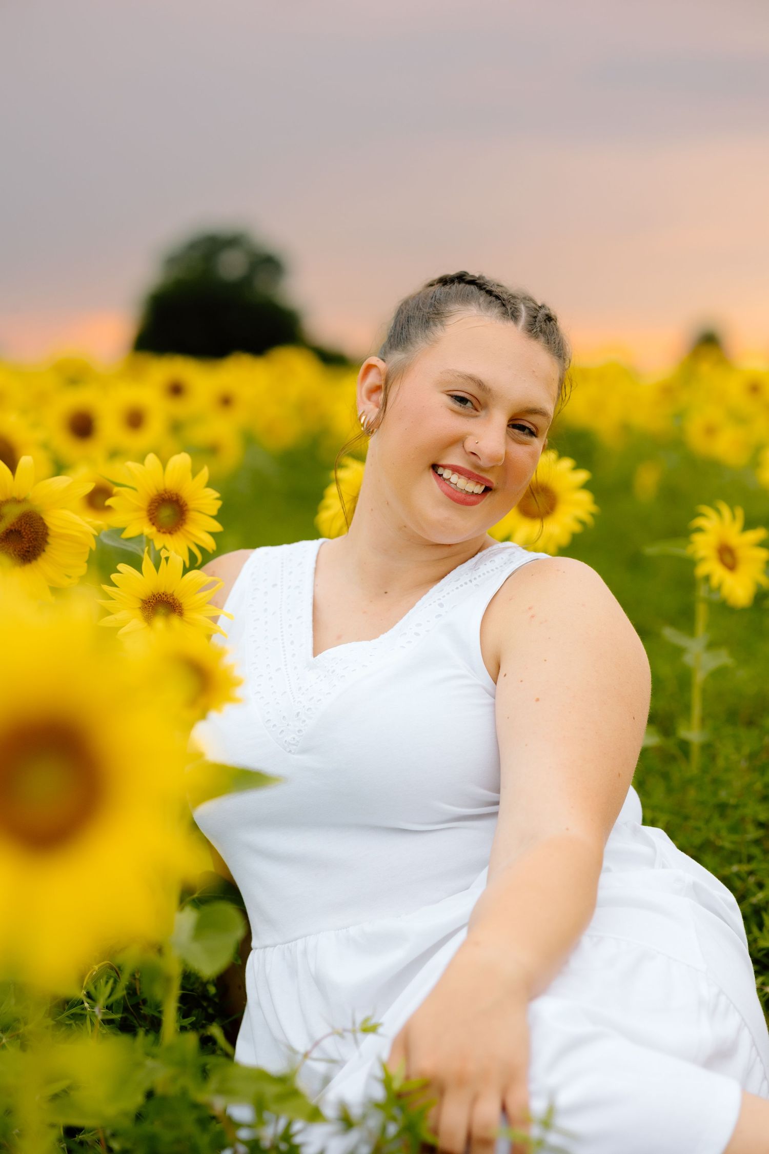 Smiling woman in white dress sitting among vibrant yellow sunflowers in a field at sunset.