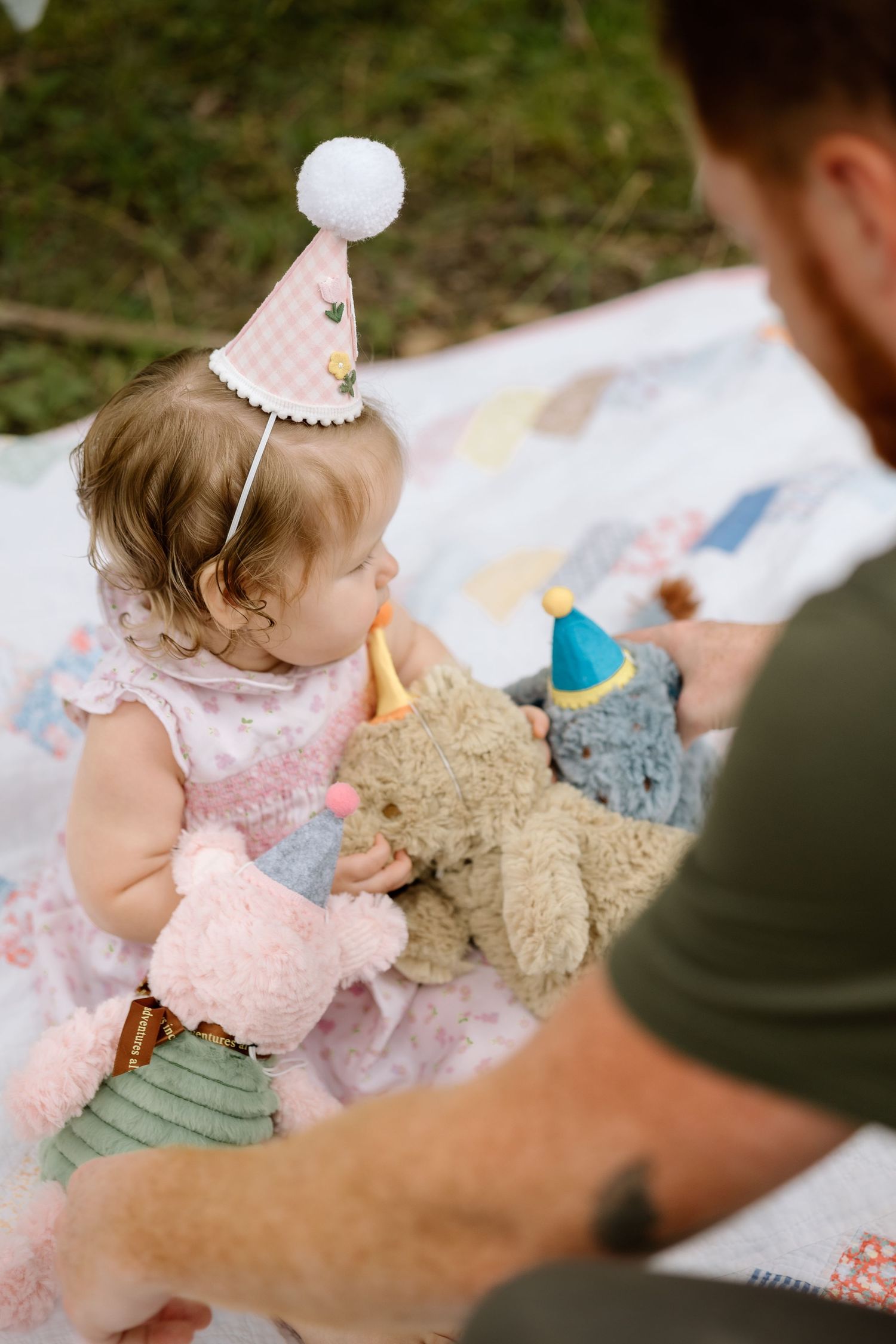 Baby girl in pink birthday hat holds stuffed animals with dad on a blanket outdoors.