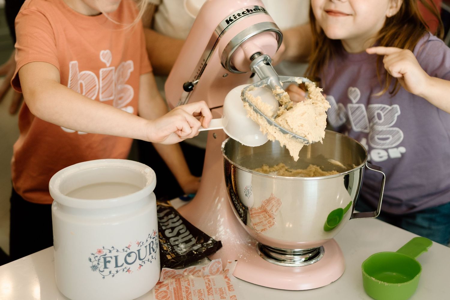 Children reaching into a pink KitchenAid mixer bowl filled with cookie dough on a kitchen counter.