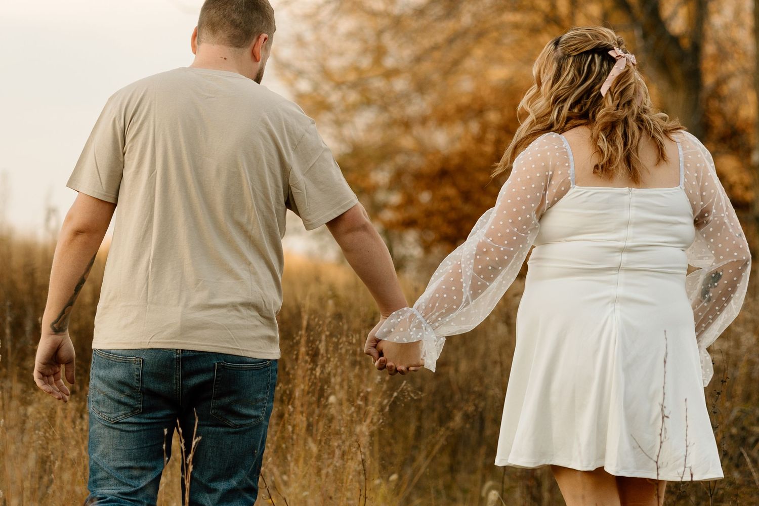 A couple walks hand in hand through an autumn field, the woman in a white lace mini dress and man in a beige shirt.