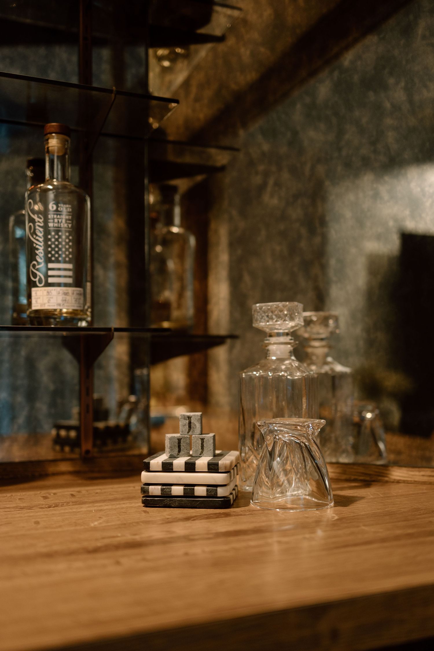 Rustic wooden bar top with crystal decanters, checkered coasters, and a whiskey bottle on dark shelves.