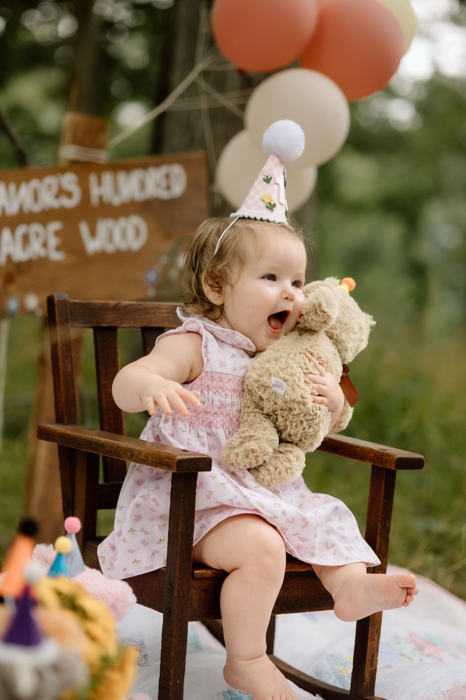 Excited birthday baby girl in party hat hugging stuffed bear in outdoor setting.