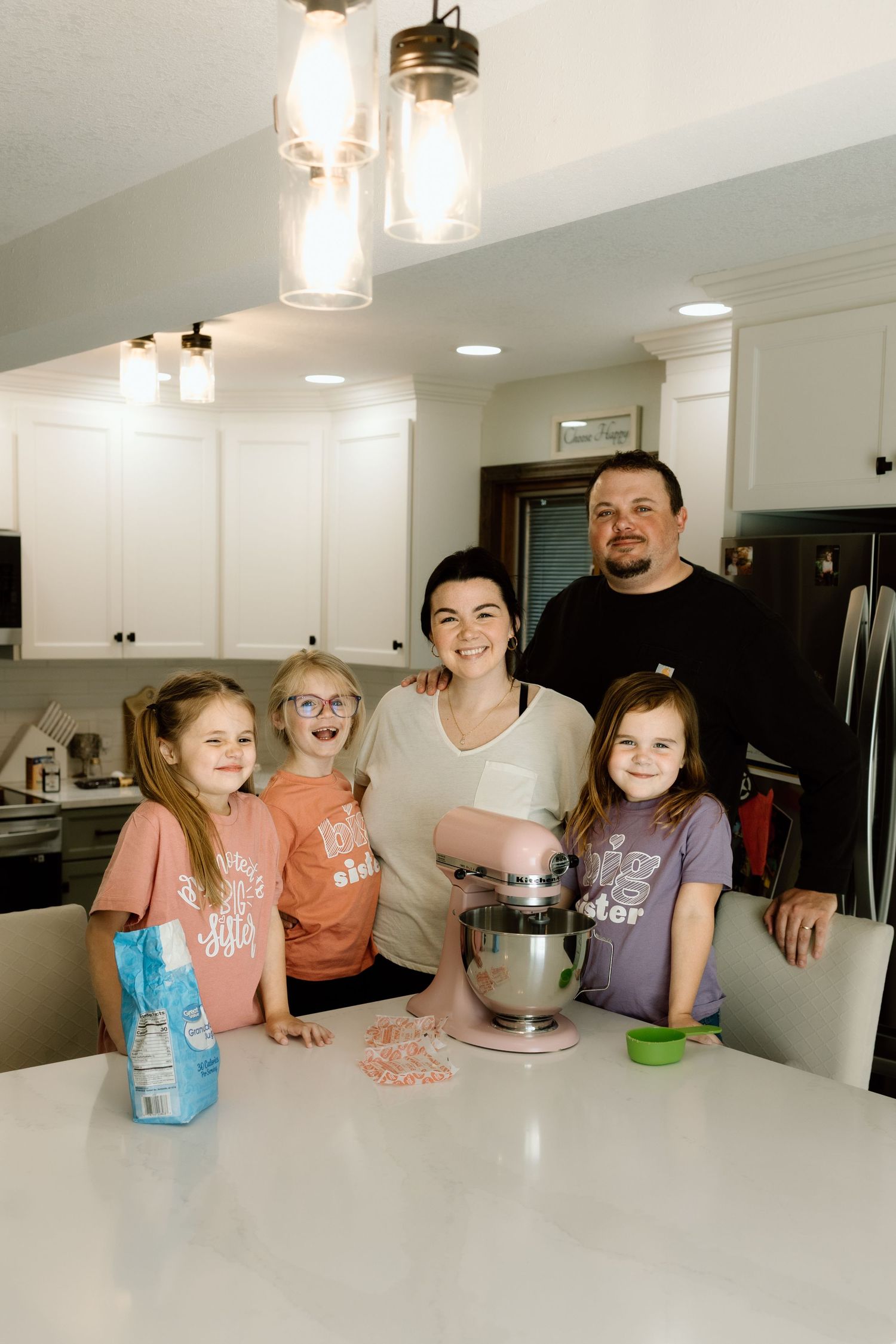 Family of five smiling together in a bright white kitchen with a pink KitchenAid mixer on the counter.