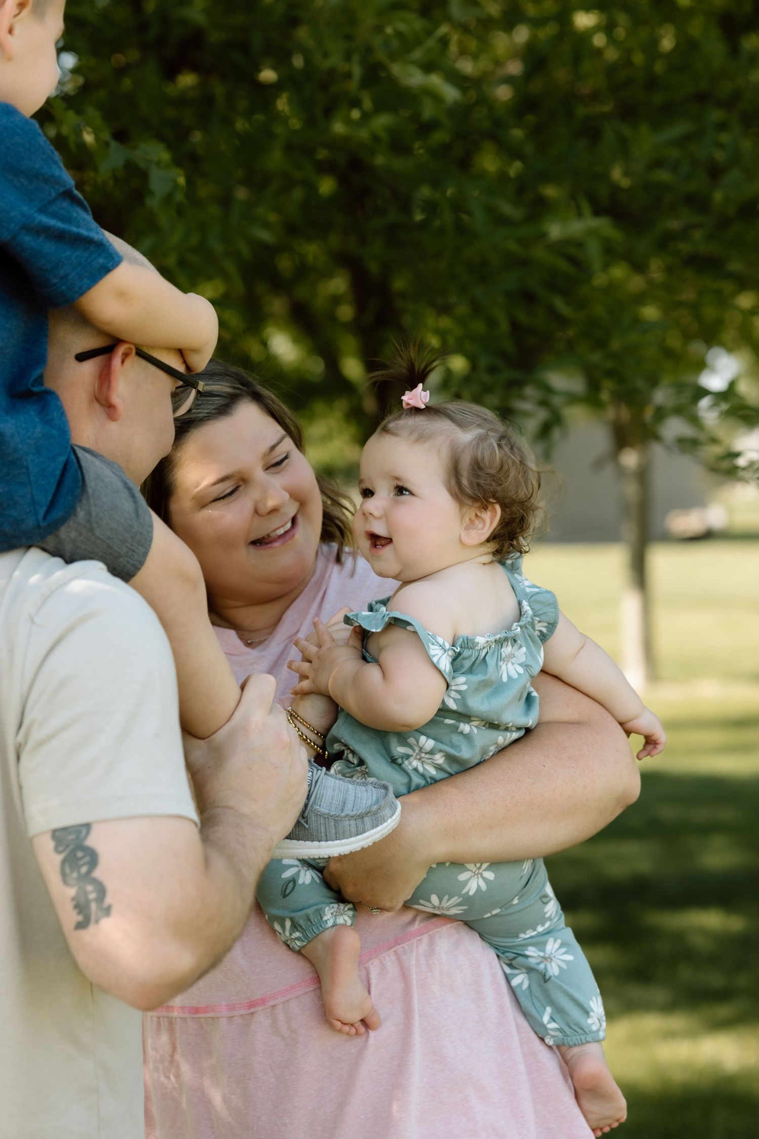 Mom laughs while holding her happy baby girl in a floral romper during a sunny outdoor family photo session.