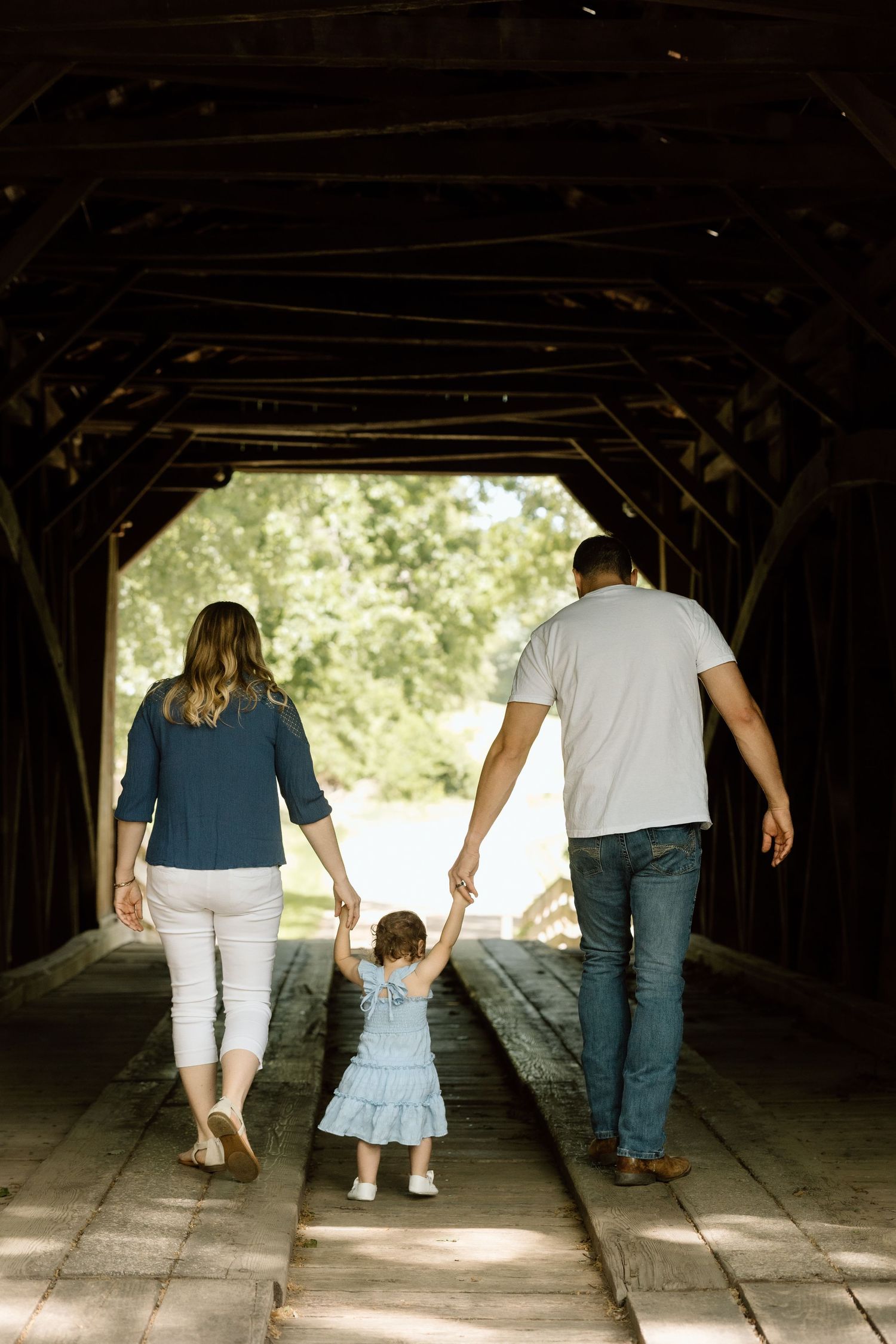 Family of three walks hand-in-hand through a historic covered wooden bridge during a family photo session.