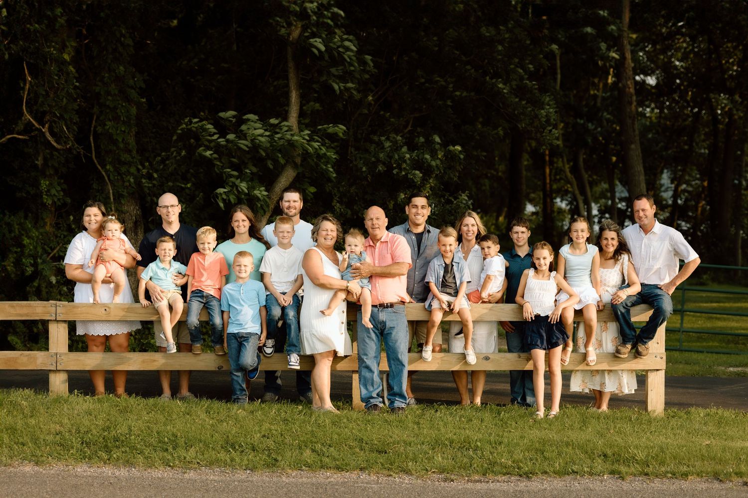 Large extended family poses together on a wooden fence in a sunny park for a group portrait outdoors.