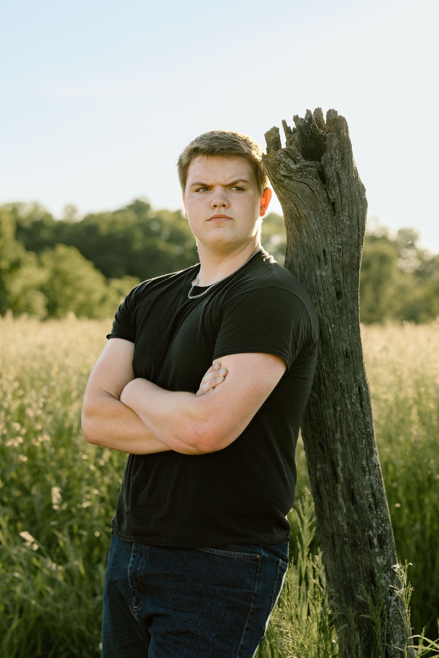 Serious teen boy with arms crossed leans against weathered wooden post in sunny wildflower field.