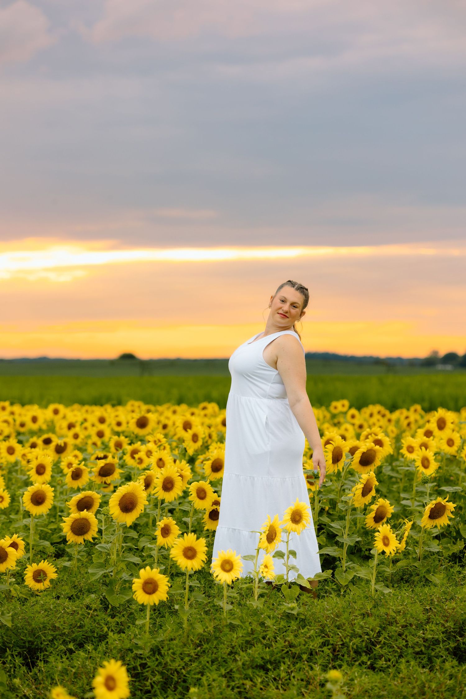 Woman in white dress standing in vast sunflower field during colorful golden sunset sky.