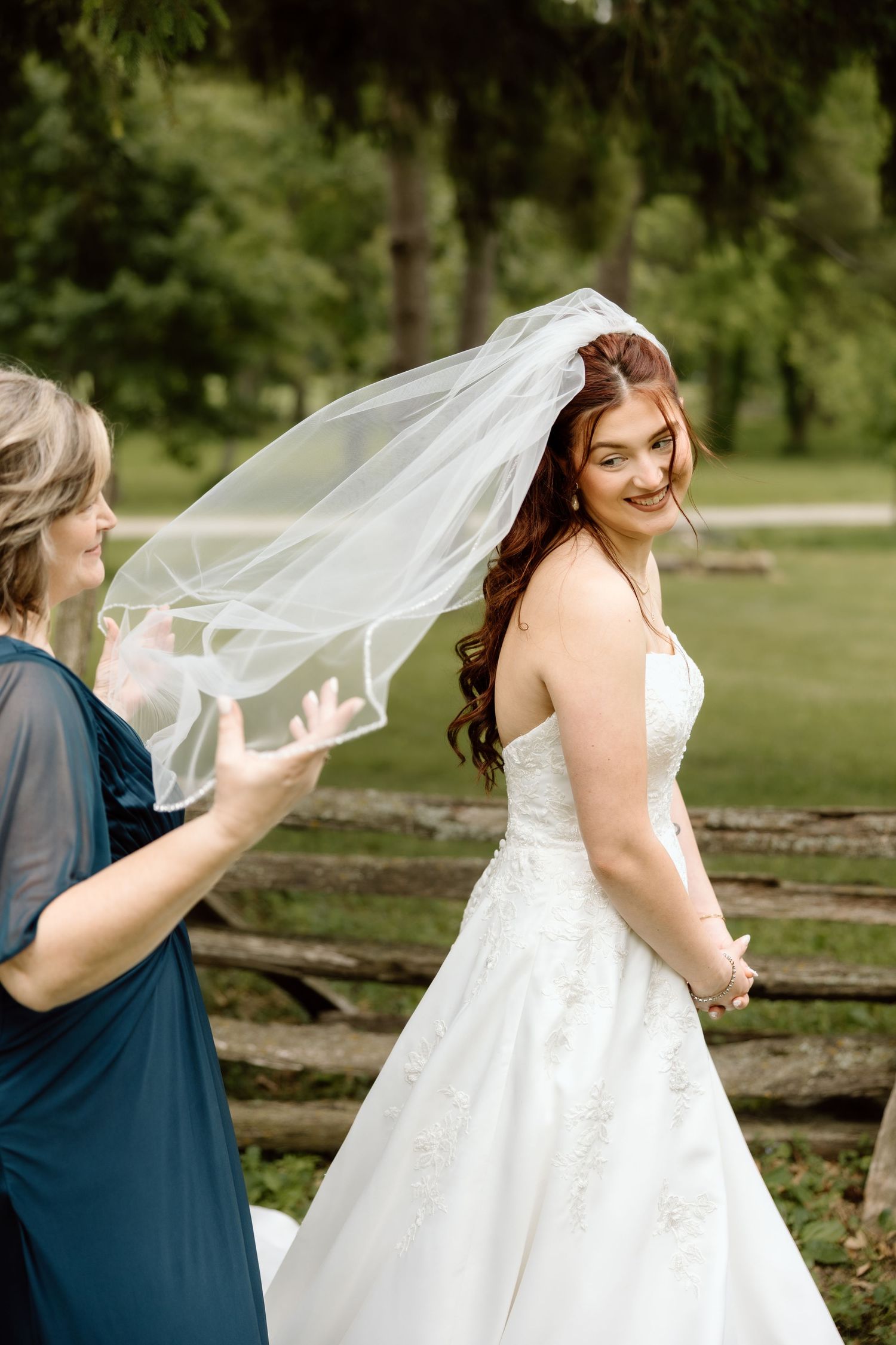 Bridesmaid adjusting bride's flowing white veil outdoors near rustic wooden fence.