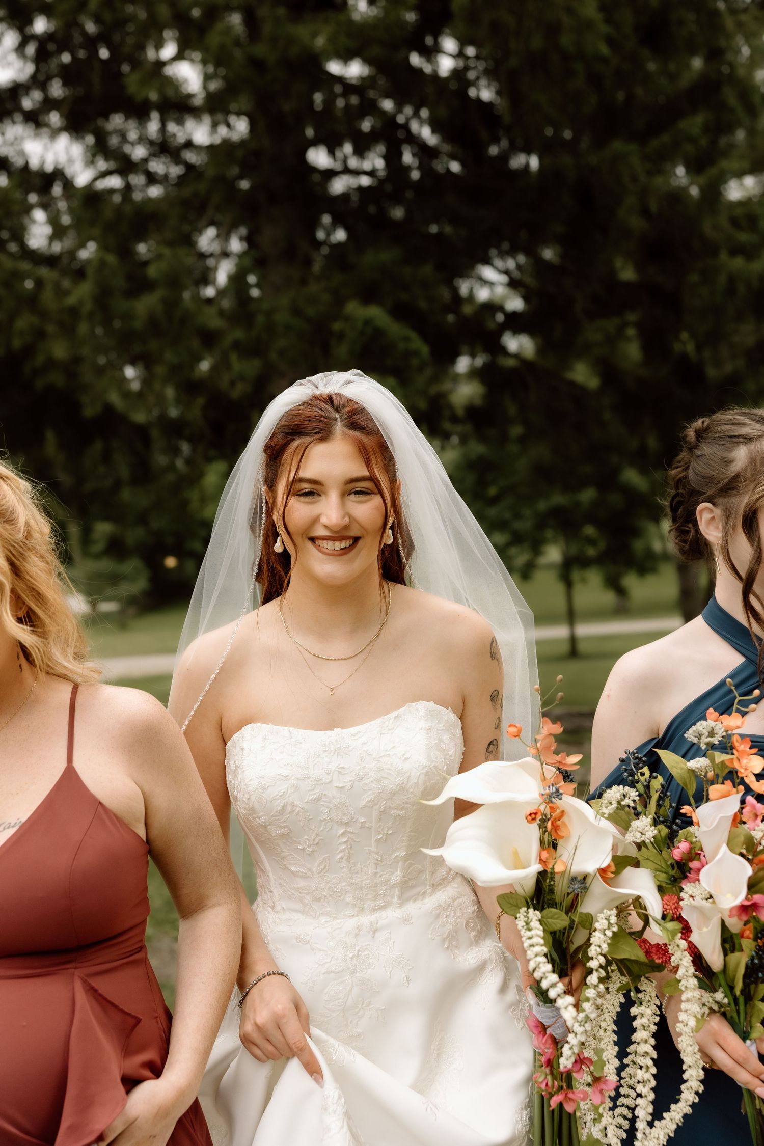 Smiling bride in white strapless lace gown wearing veil walks with bridesmaids in colorful dresses.