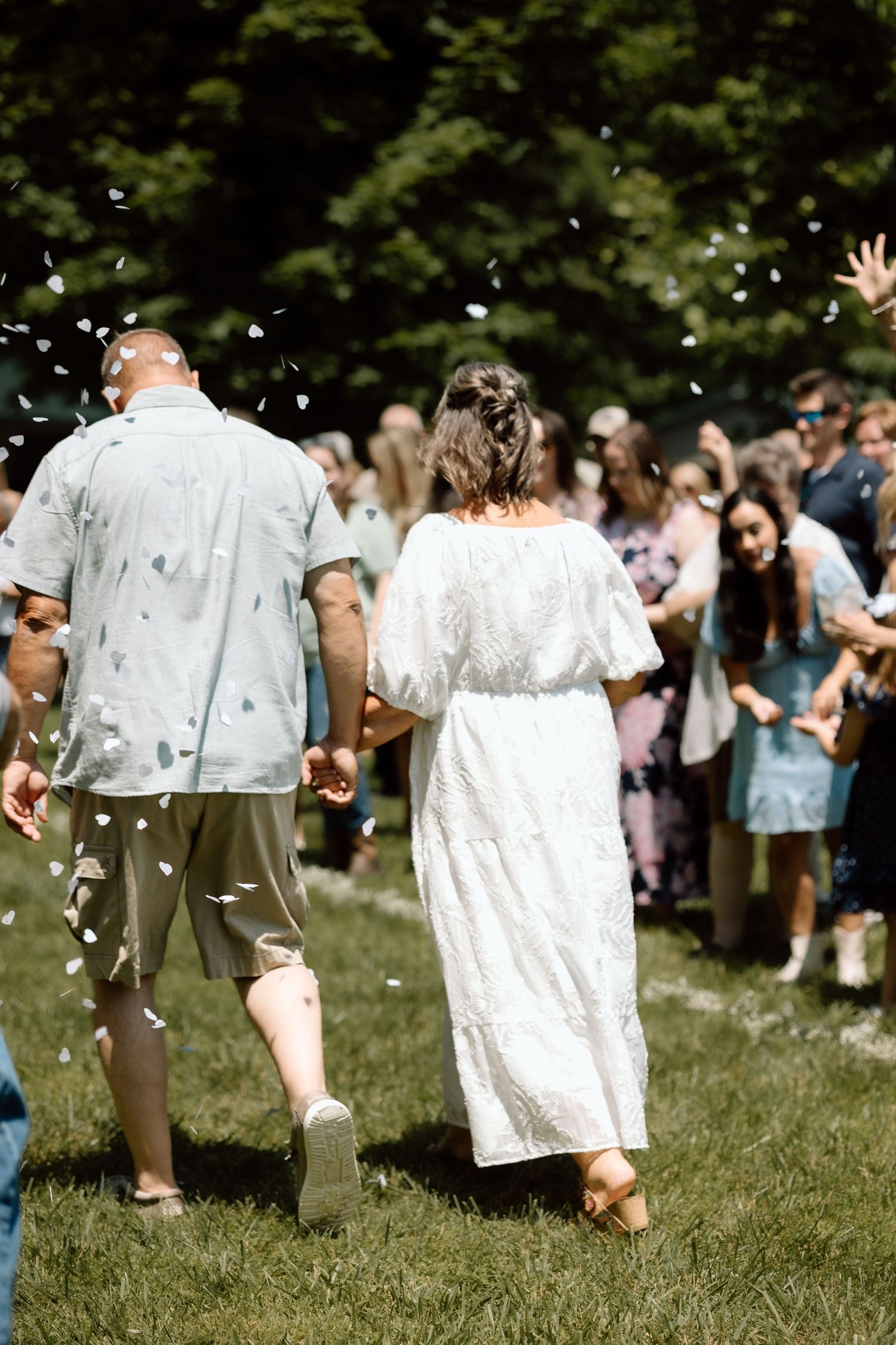 Newlywed couple walks hand-in-hand through confetti shower as guests celebrate their marriage outdoors.