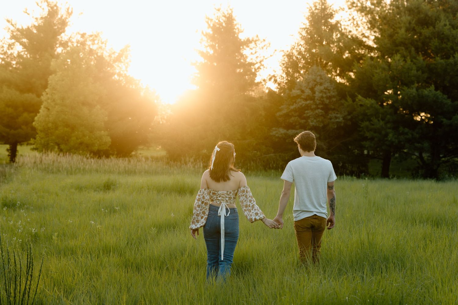 Couple holding hands walking toward sunset in lush green meadow surrounded by tall trees.