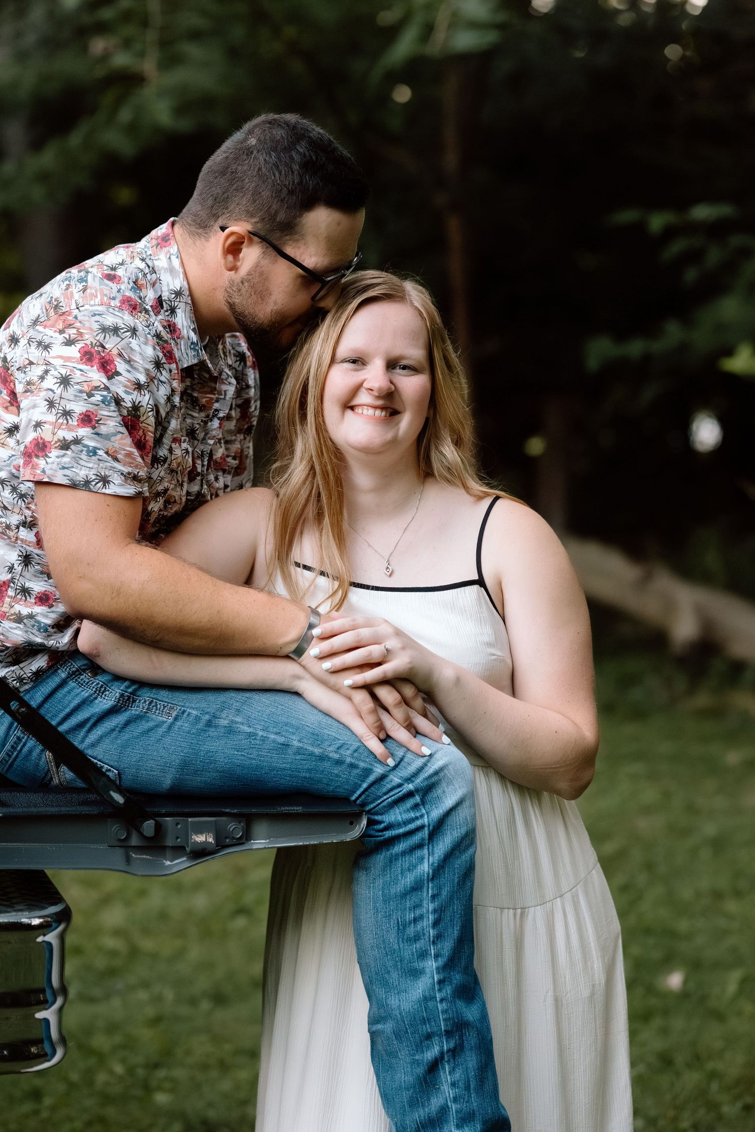 A smiling woman in a white dress sits as her partner kisses her cheek while perched on a truck tailgate outdoors.