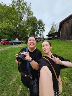 Two women smile for a selfie outdoors near a rustic barn, one holding a camera while the other flashes a peace sign.