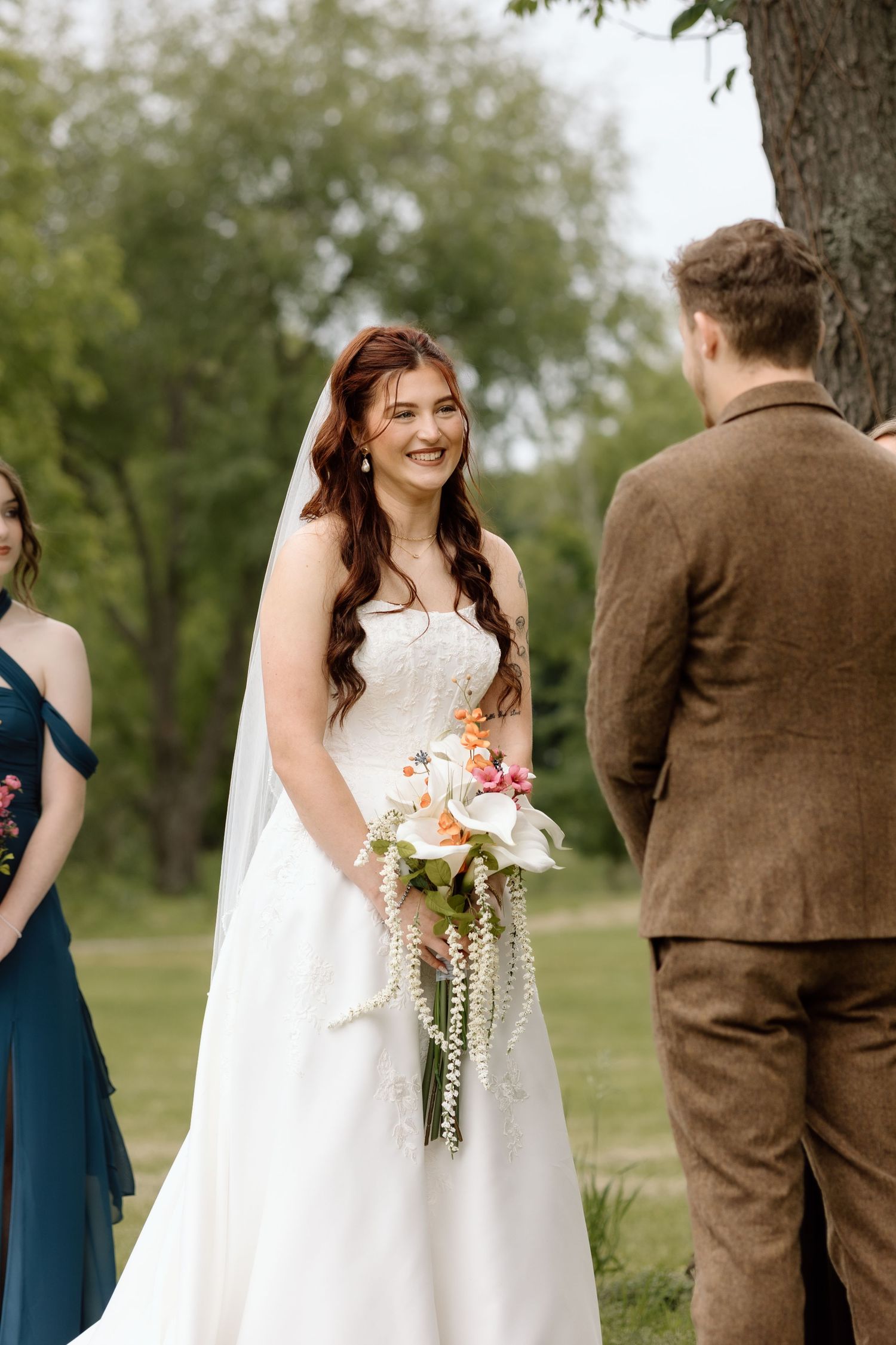 Radiant bride in strapless white gown holds colorful cascading bouquet while smiling at groom during outdoor ceremony.