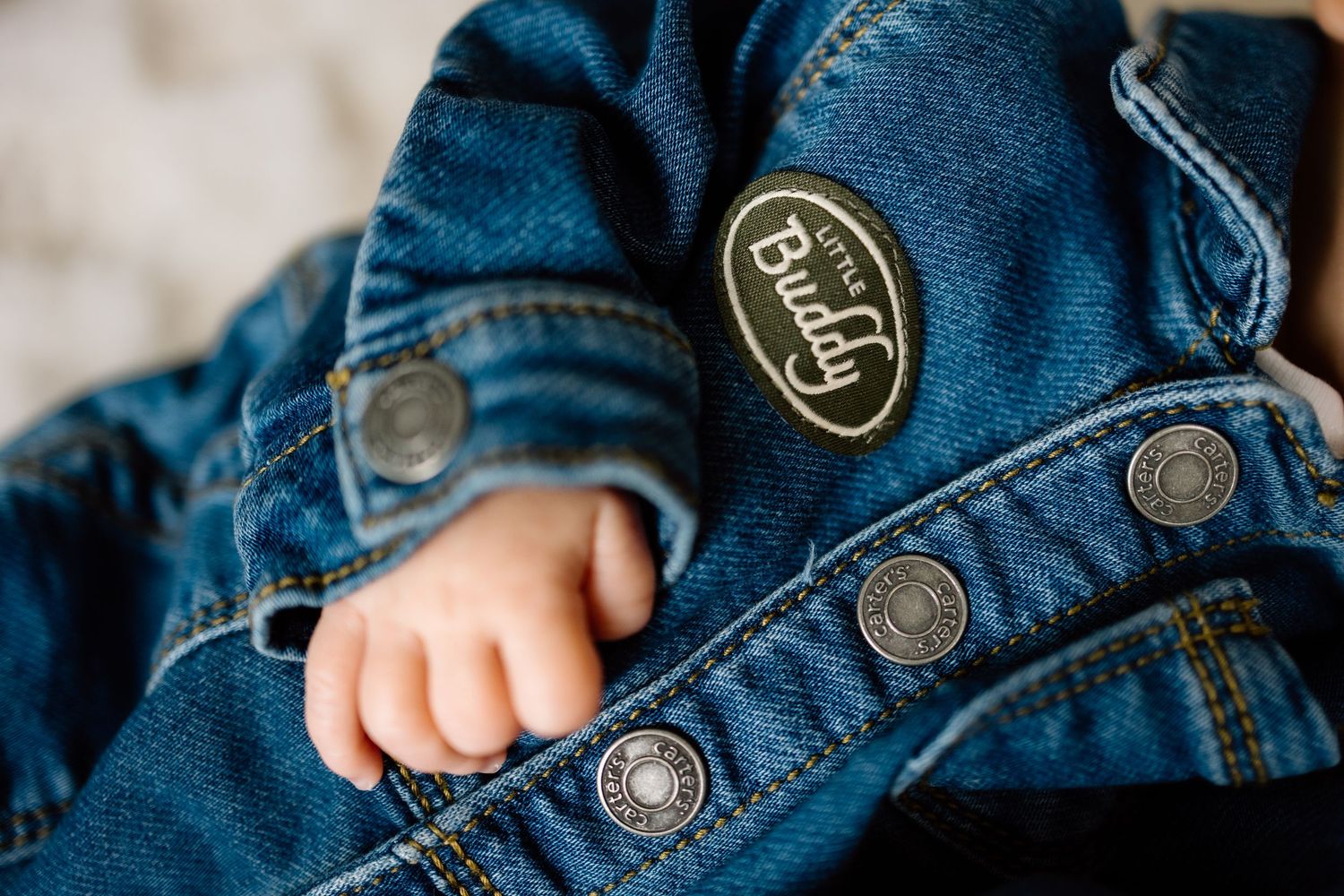 Close-up of a baby hand near Little Buddy patch on a denim jacket with snap buttons.