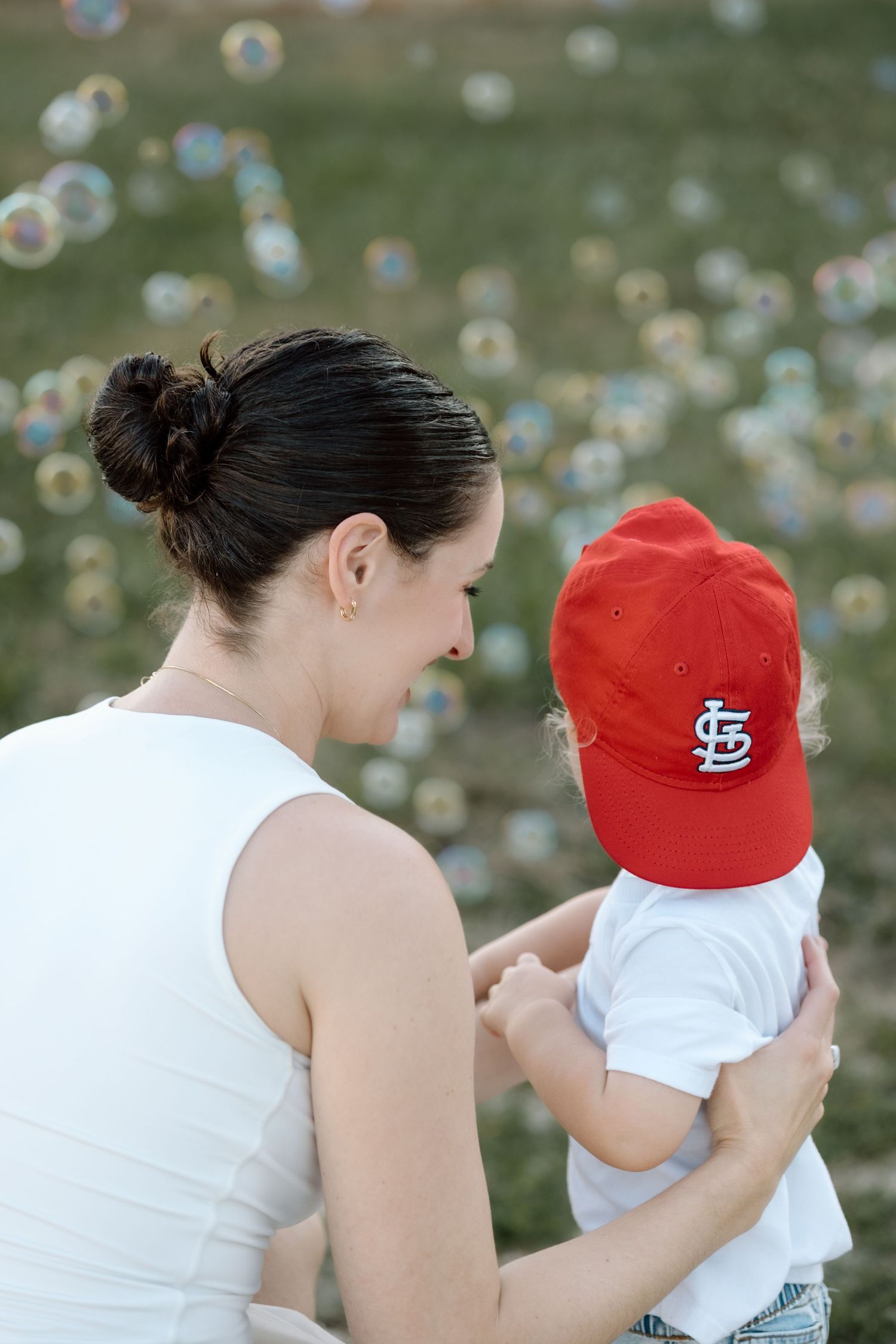 Mother in white top holding her baby boy in red Cardinals baseball cap outdoors.
