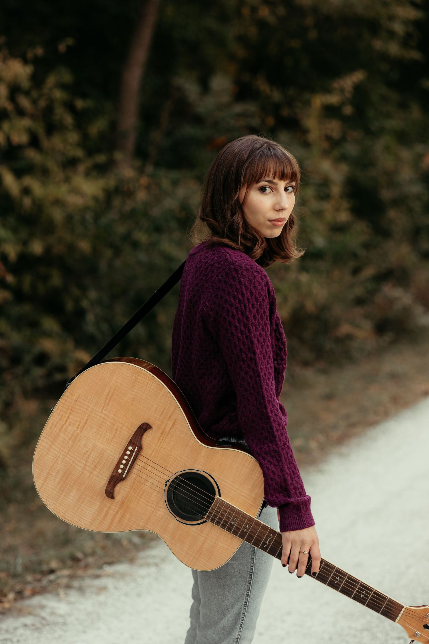 Young woman in purple sweater carries acoustic guitar on a tree-lined gravel path.
