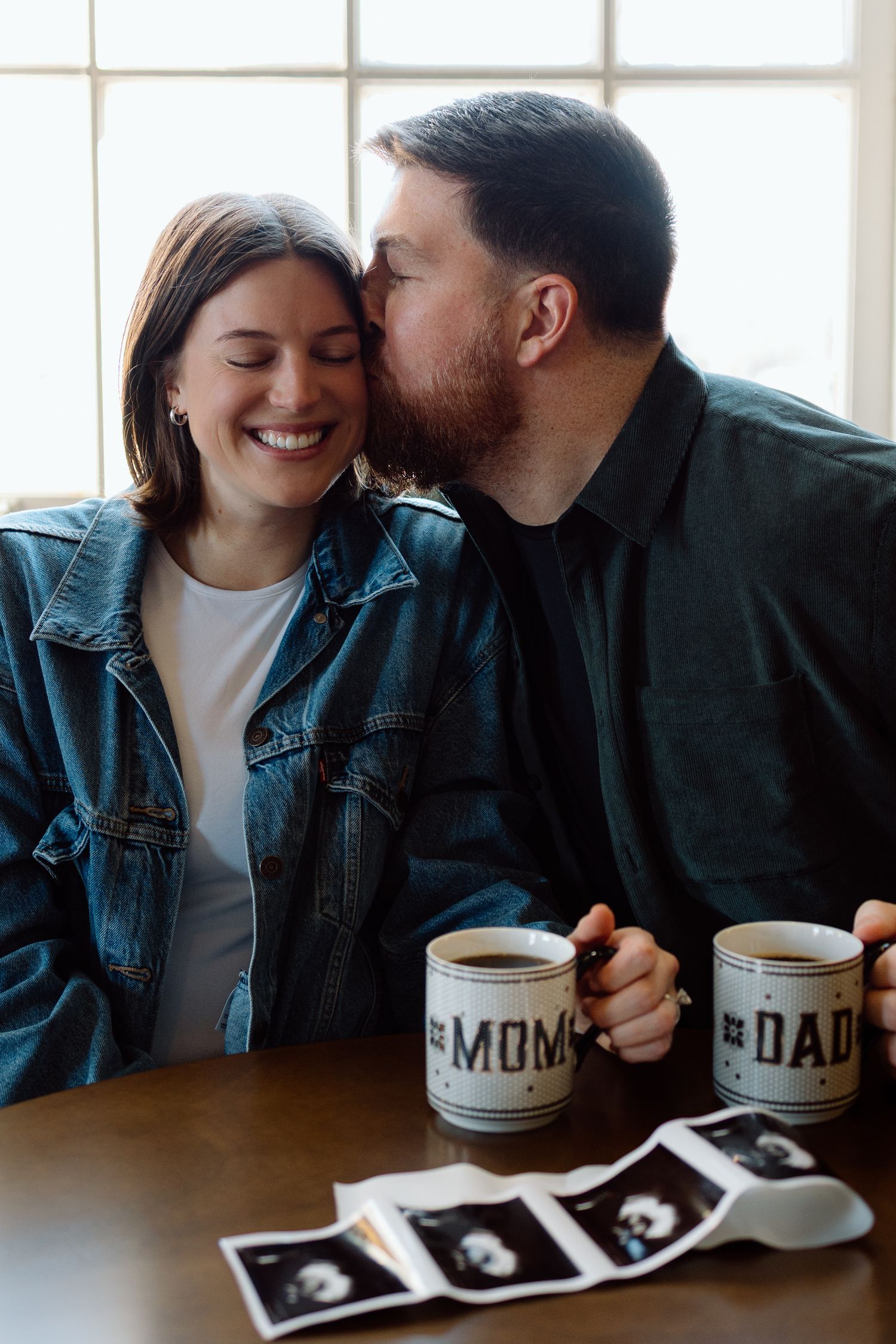 Couple sharing joyful moment with Mom and Dad mugs and ultrasound photos on table.