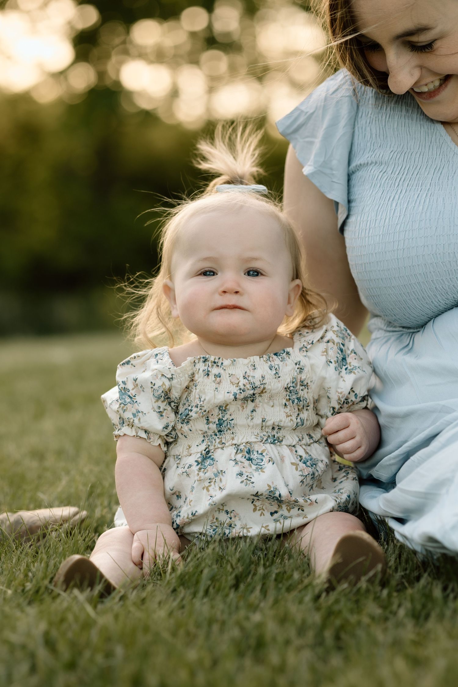 An adorable baby girl in a floral dress sits on green grass beside her mother during a sunset photo session.