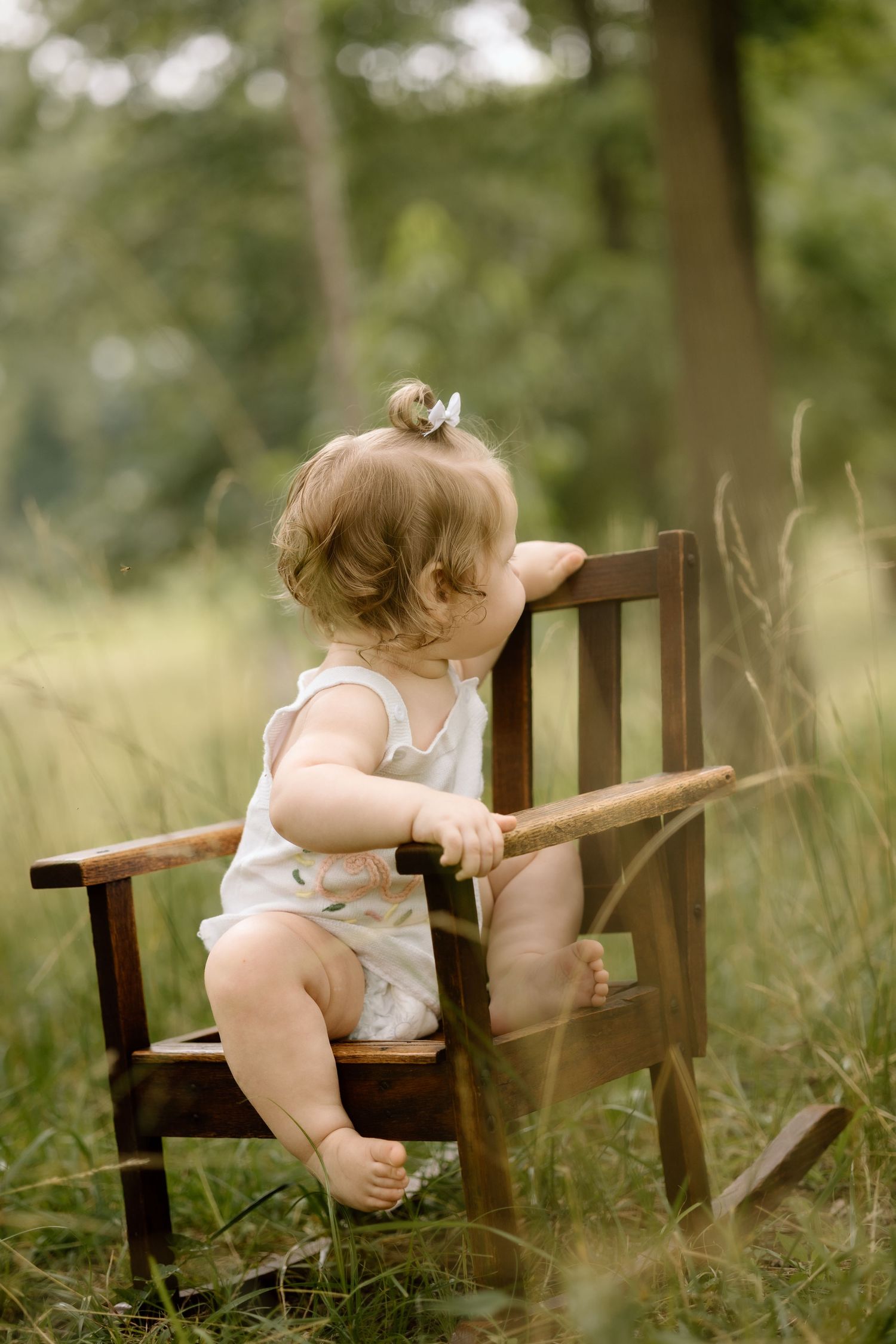 Adorable baby in white romper climbing on small wooden chair in lush green outdoor setting.