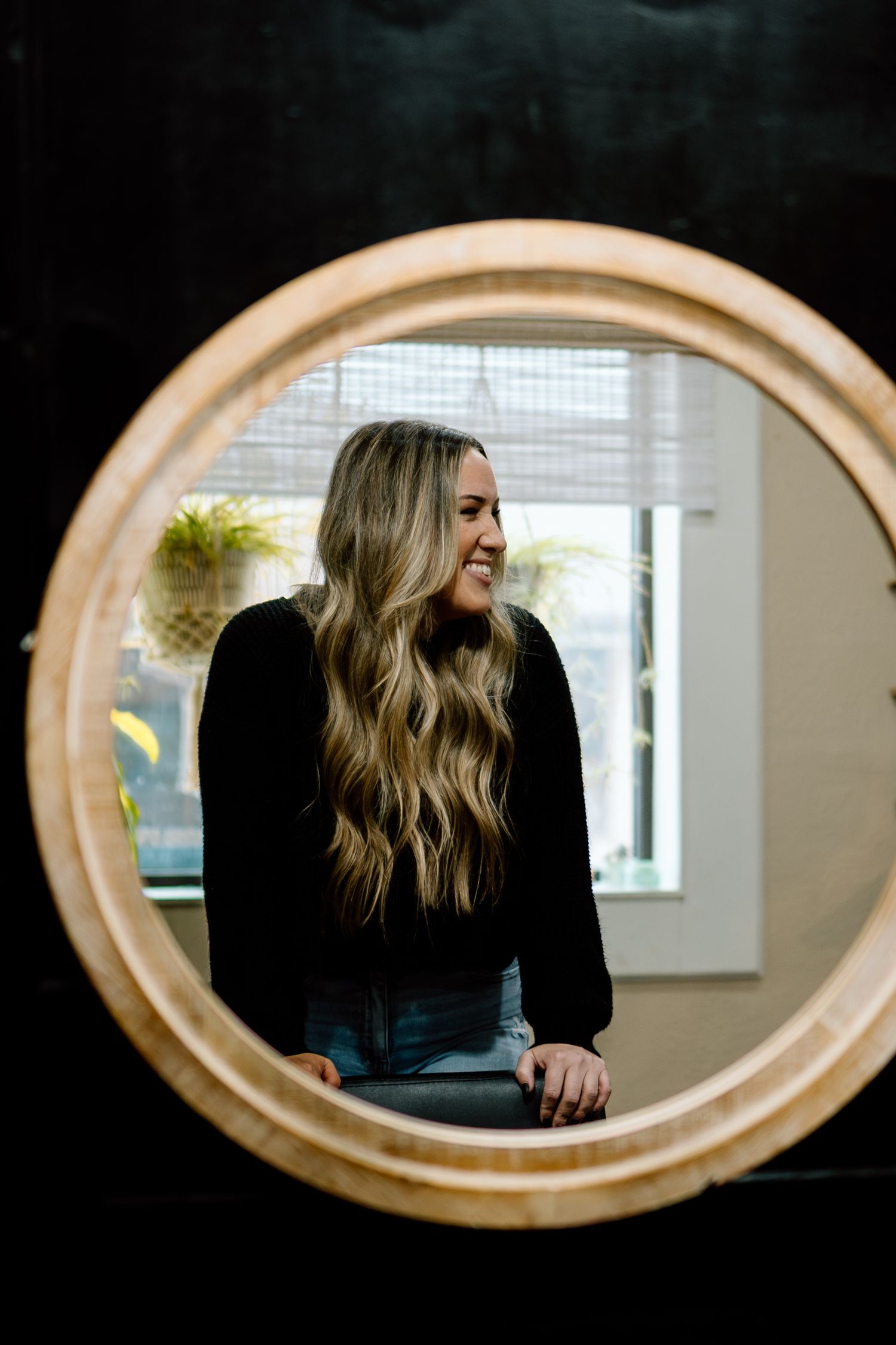 A smiling blonde woman with long wavy hair reflected in a round wooden framed mirror.