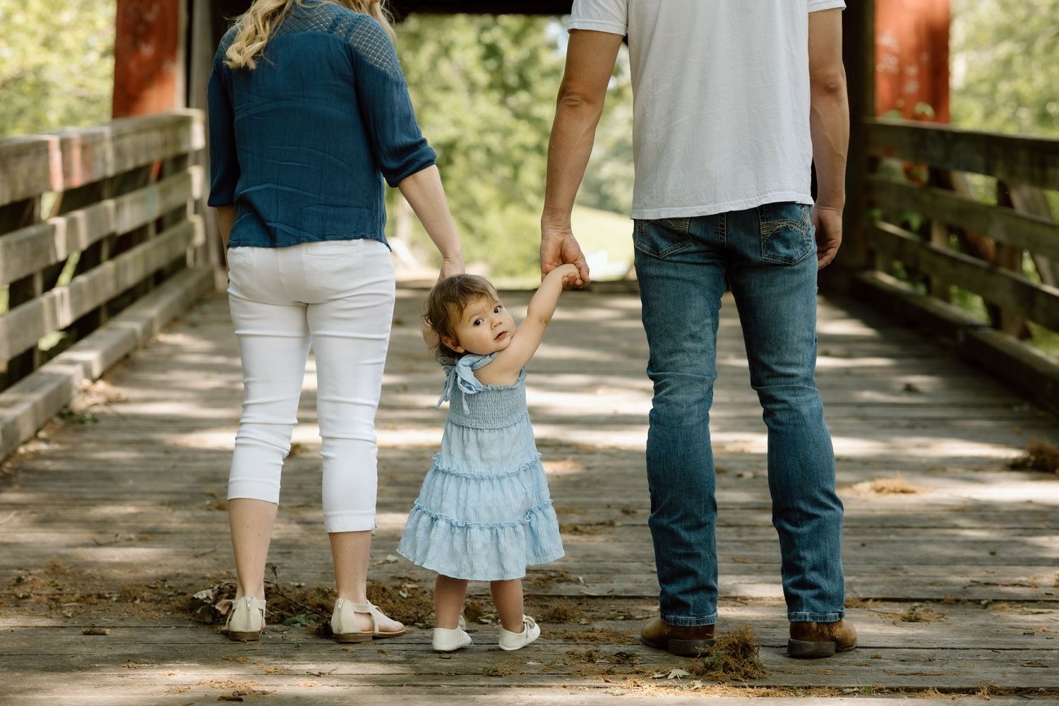 Parents and toddler daughter in blue dress walk hand in hand across a wooden bridge during family photo session.