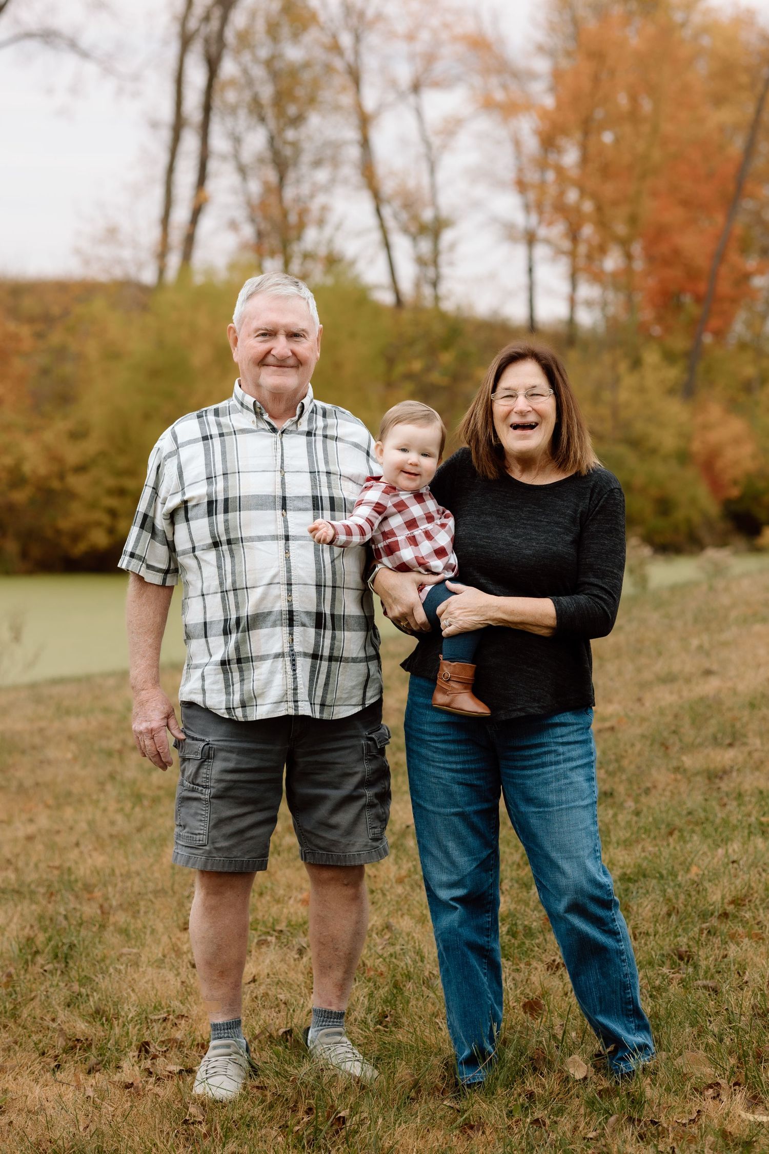 Grandparents smiling proudly while holding a baby in a plaid shirt during an outdoor fall photo session.