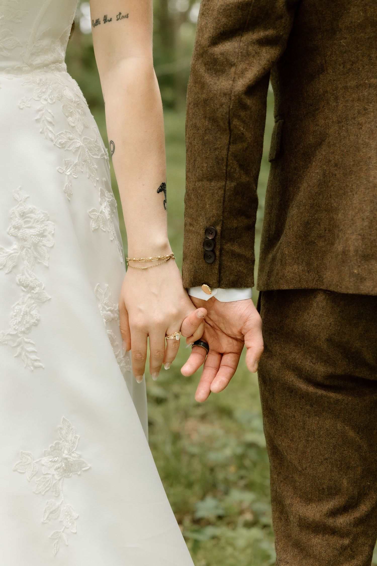 Close-up of bride and groom holding hands, bride wearing gold bracelet and ring in lace wedding dress.