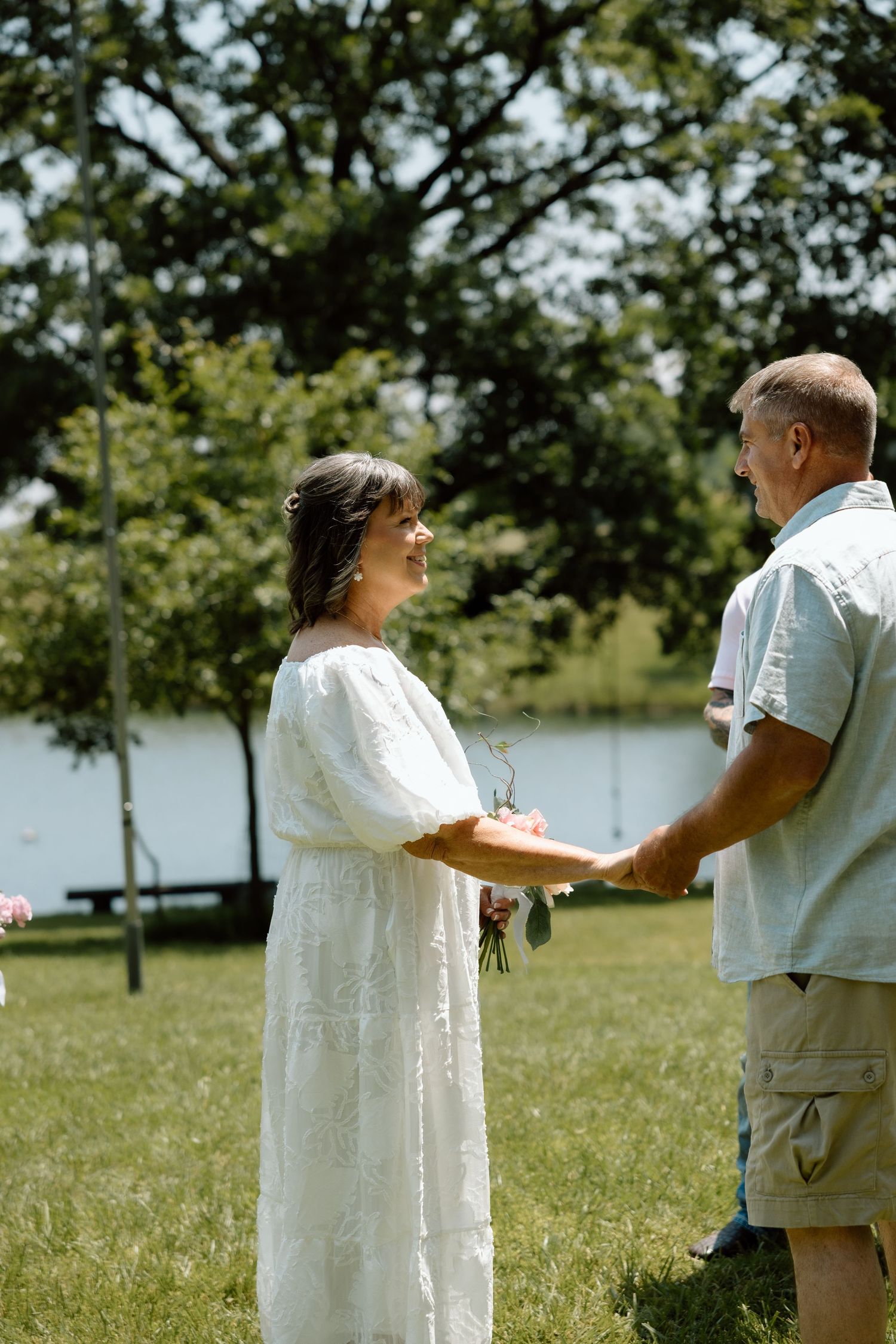 Bride in white lace dress holds bouquet while exchanging vows with groom outdoors near a lake.