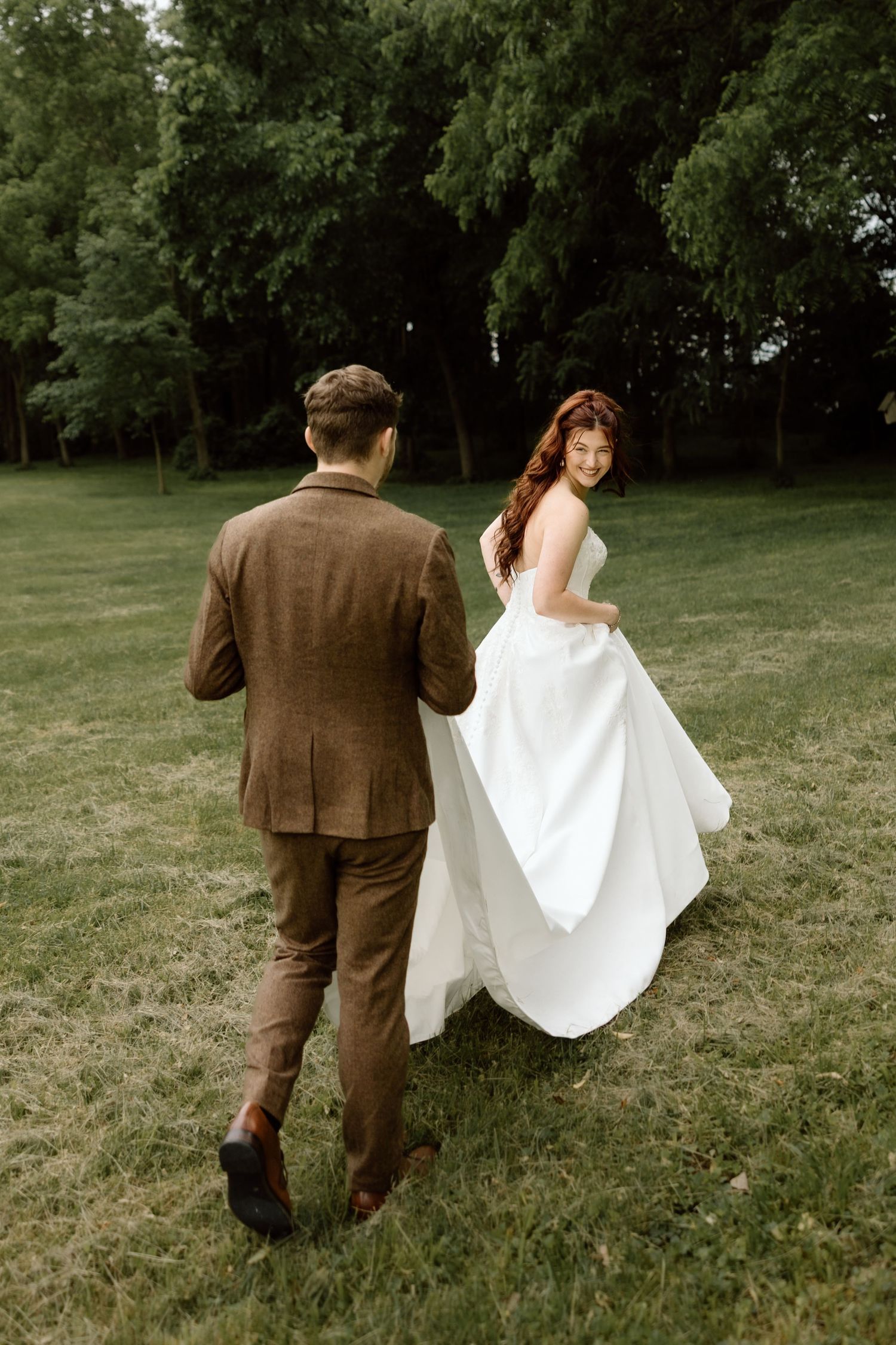 Bride in flowing white gown glancing back at groom while walking across a lush green lawn.