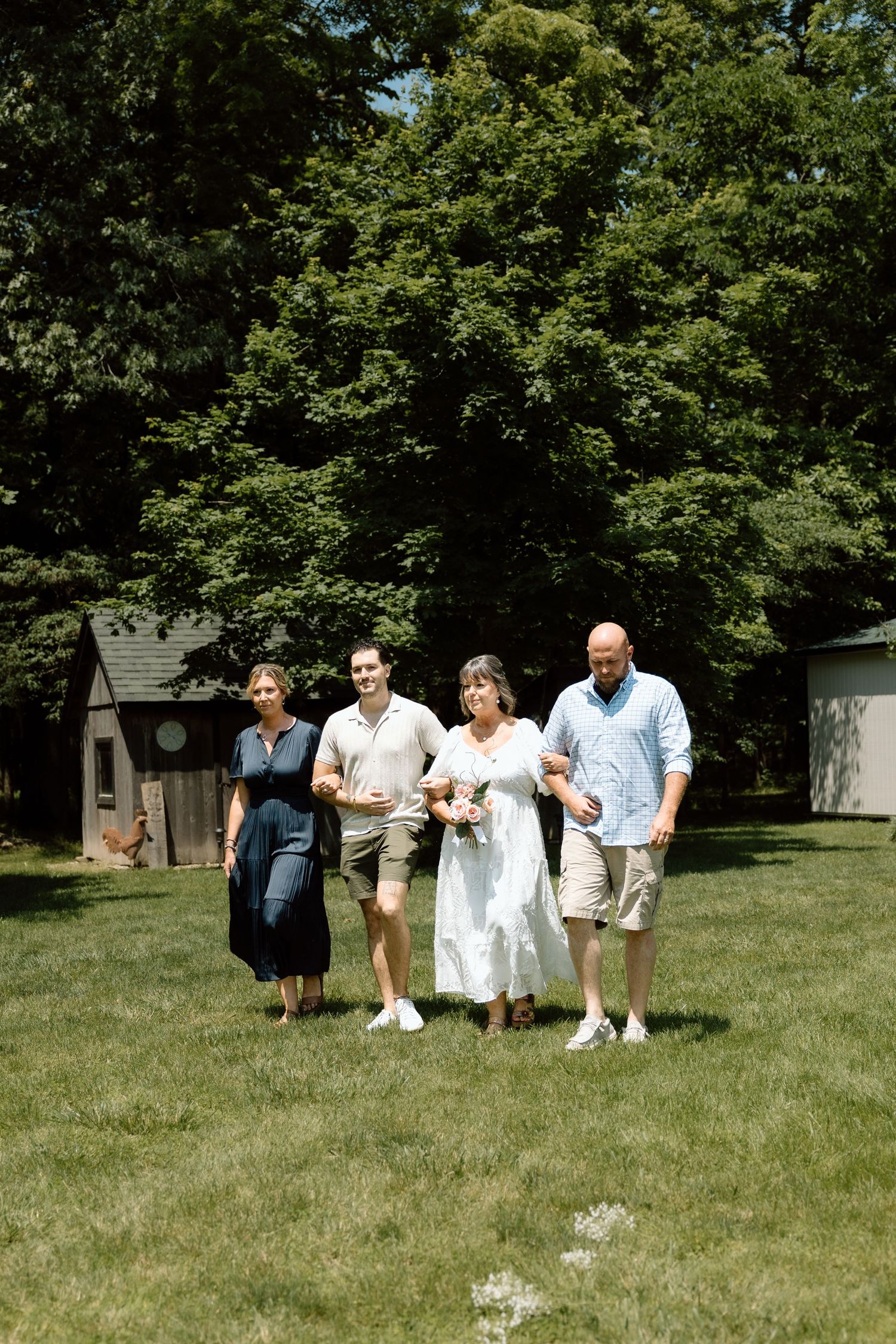 Wedding party of four walking together across green lawn with trees and small shed in background.