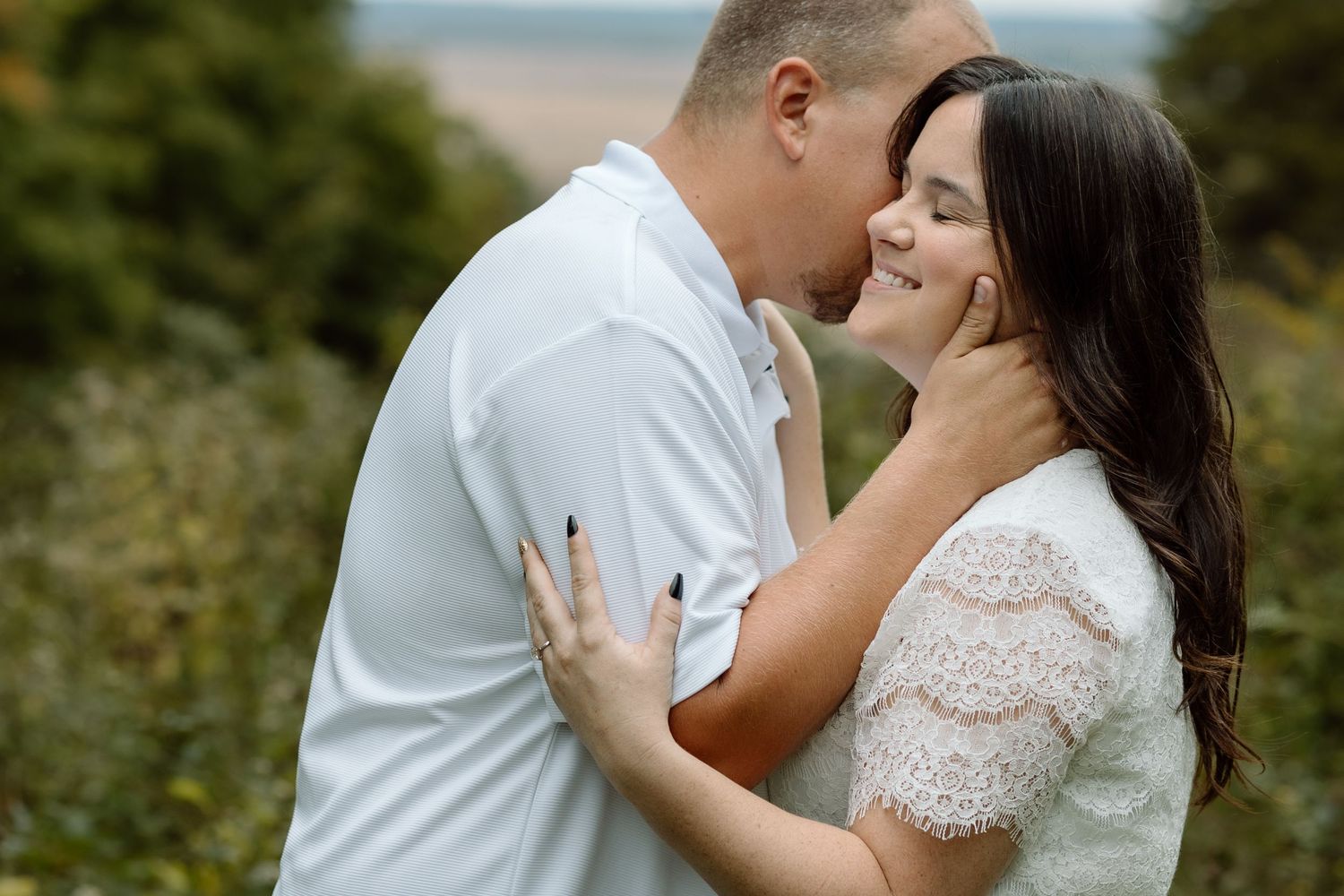 Couple sharing a tender almost-kiss moment outdoors, woman smiling as man holds her face gently near the coast.