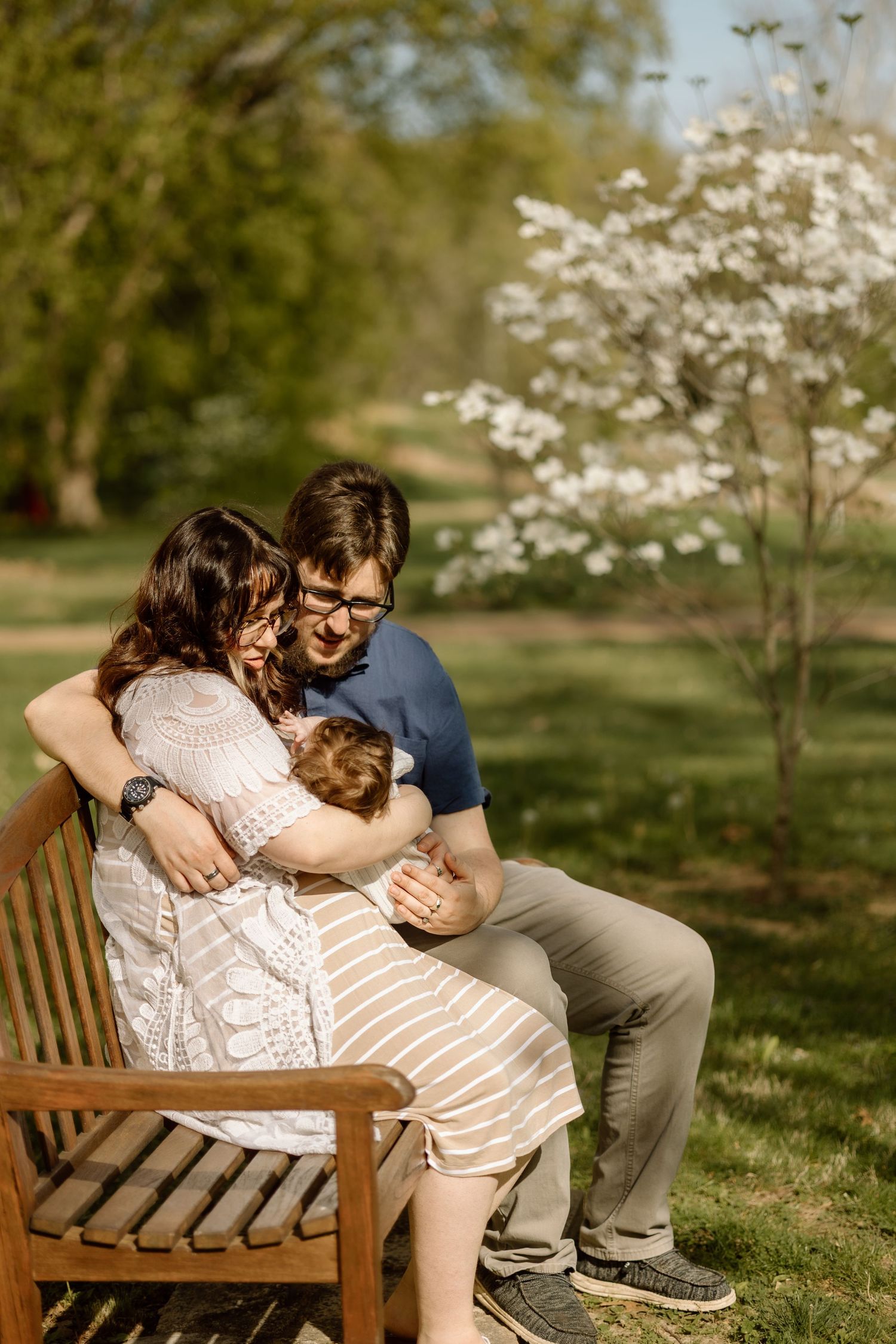 Couple sits on wooden bench embracing their newborn baby near blooming white dogwood tree in spring park.