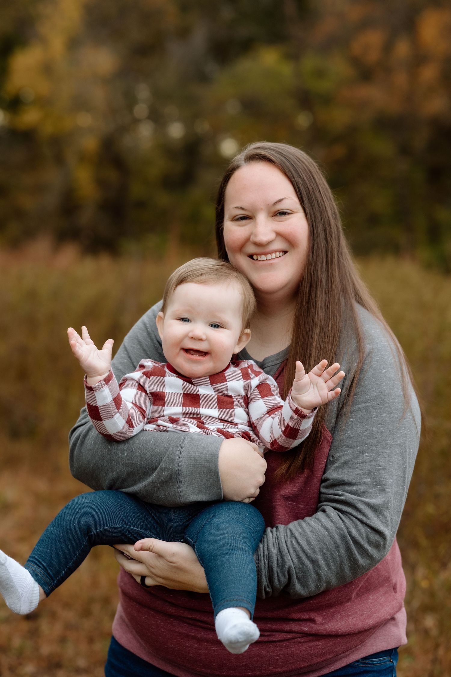 Smiling brunette woman holding a cheerful baby with raised hands during an outdoor fall family portrait.