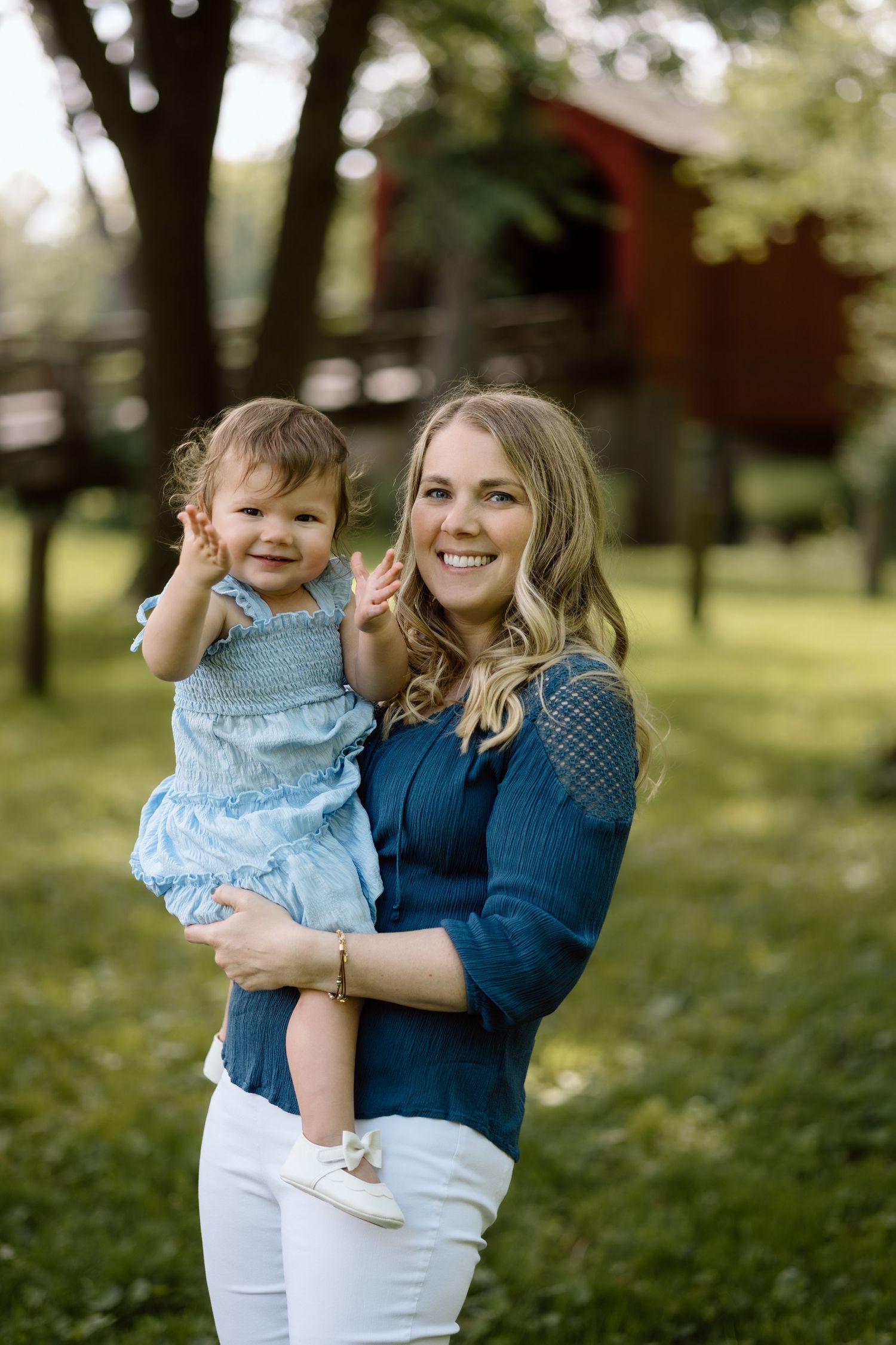 Smiling blonde mother holds her happy baby girl in a blue dress during an outdoor portrait session near red barn.