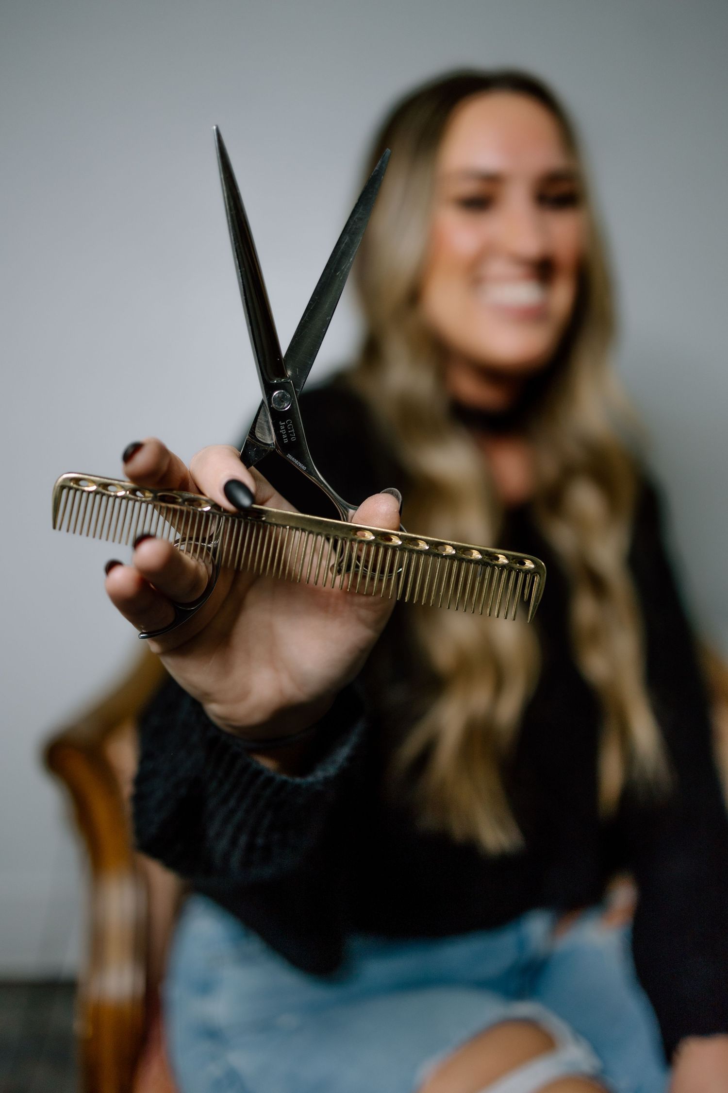 A smiling blonde hairstylist holds scissors and a comb toward the camera in a salon.