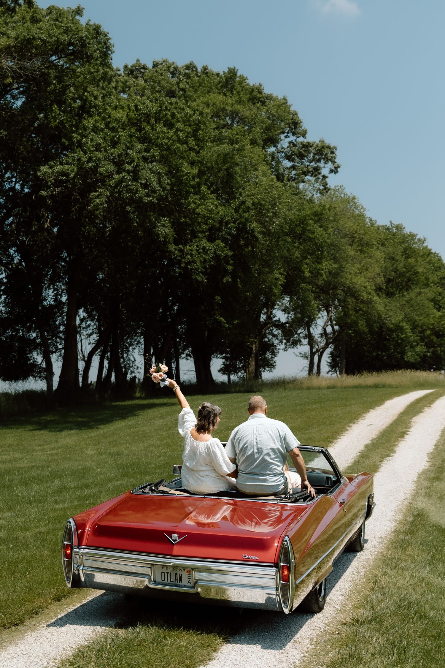 Bride with raised arm and groom sitting on trunk of red classic convertible on scenic country road.
