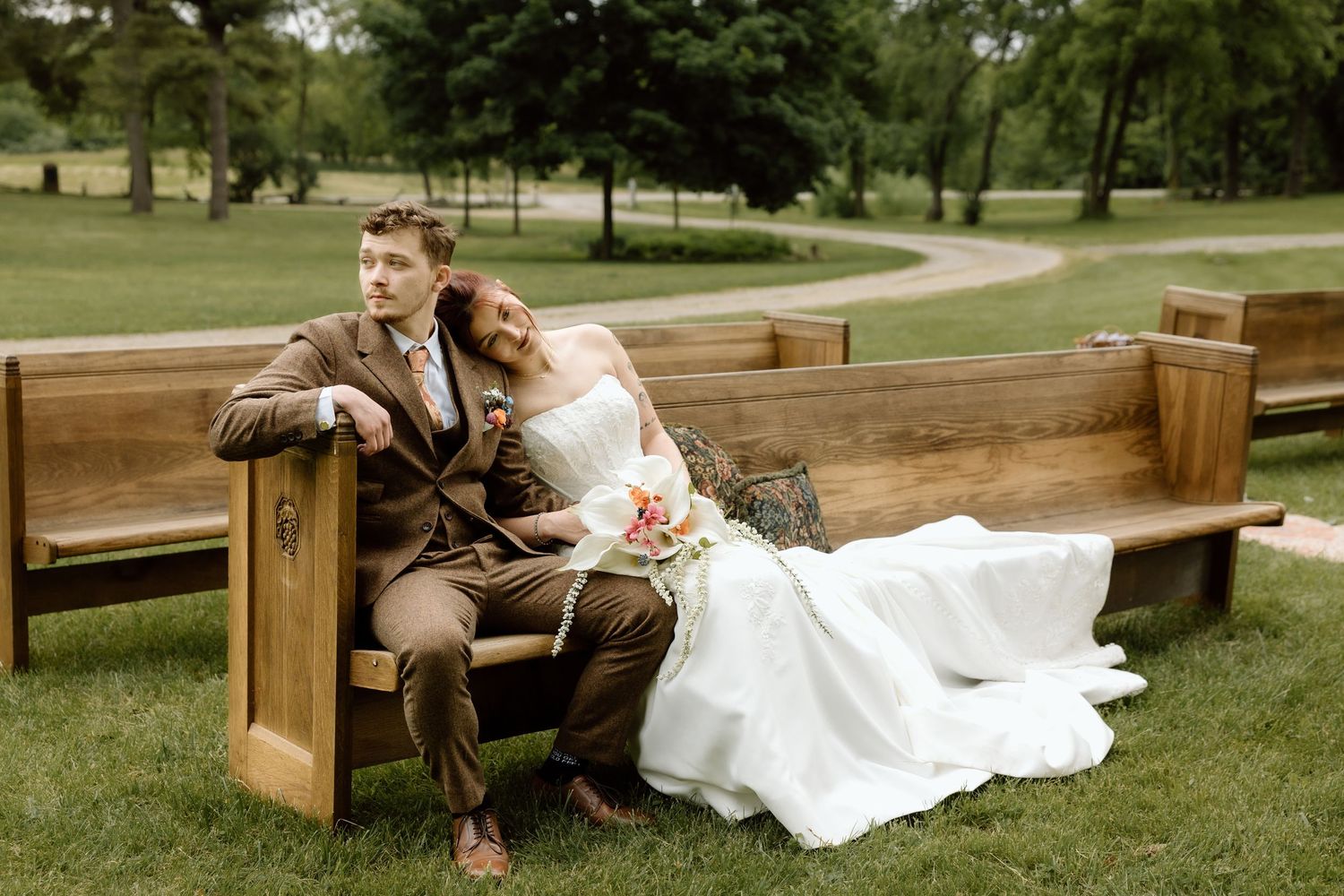 Bride and groom sitting on a wooden pew bench in a lush green park during wedding portrait session.
