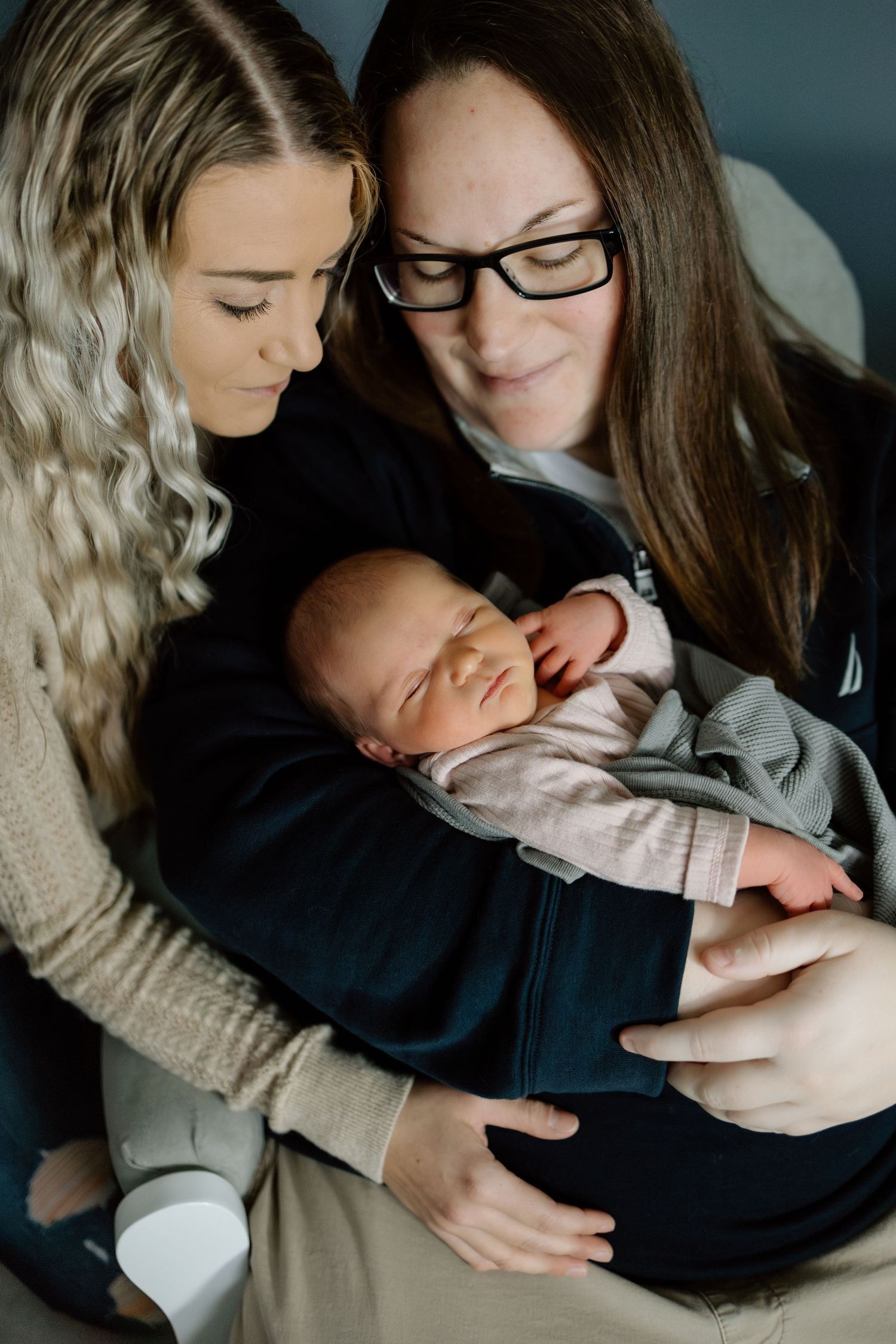 Two women lovingly gazing at a sleeping newborn baby cradled in their arms together.